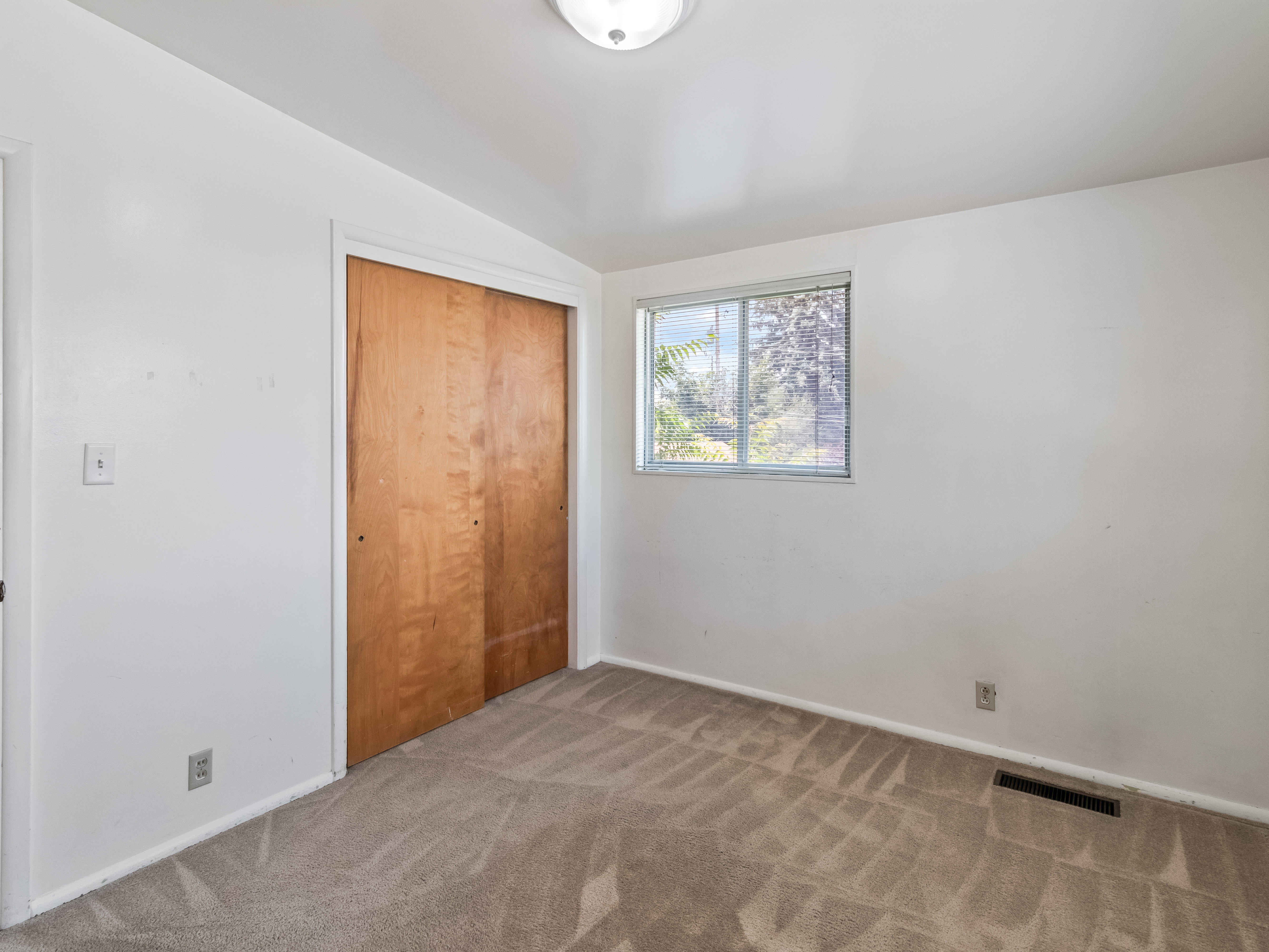 Carpeted room featuring a window with horizontal blinds, light wood-finish bi-fold closet doors, and white walls