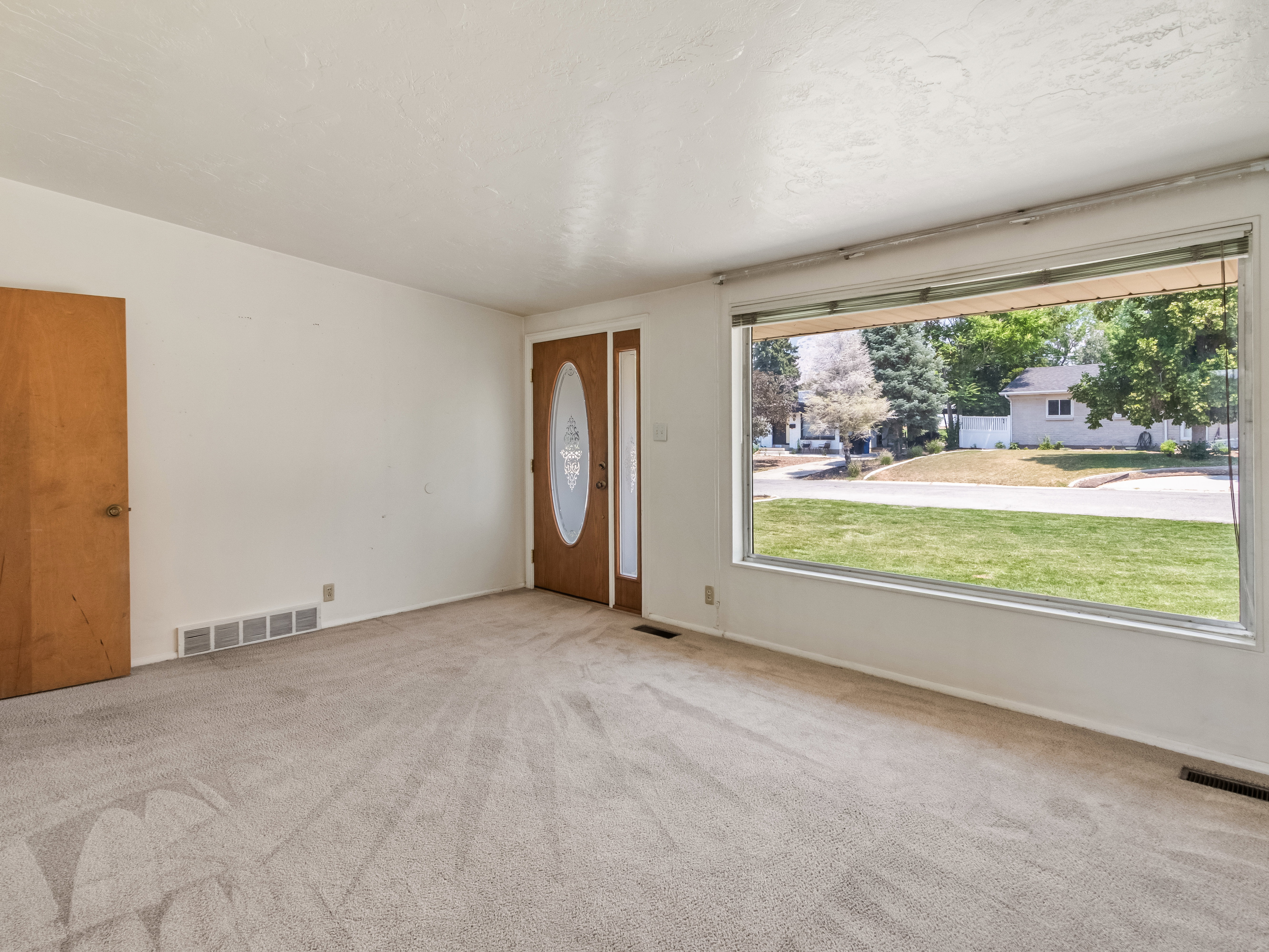 Living area featuring a large picture window, light-toned carpeting,  and a wood-panel entry door with decorative glass