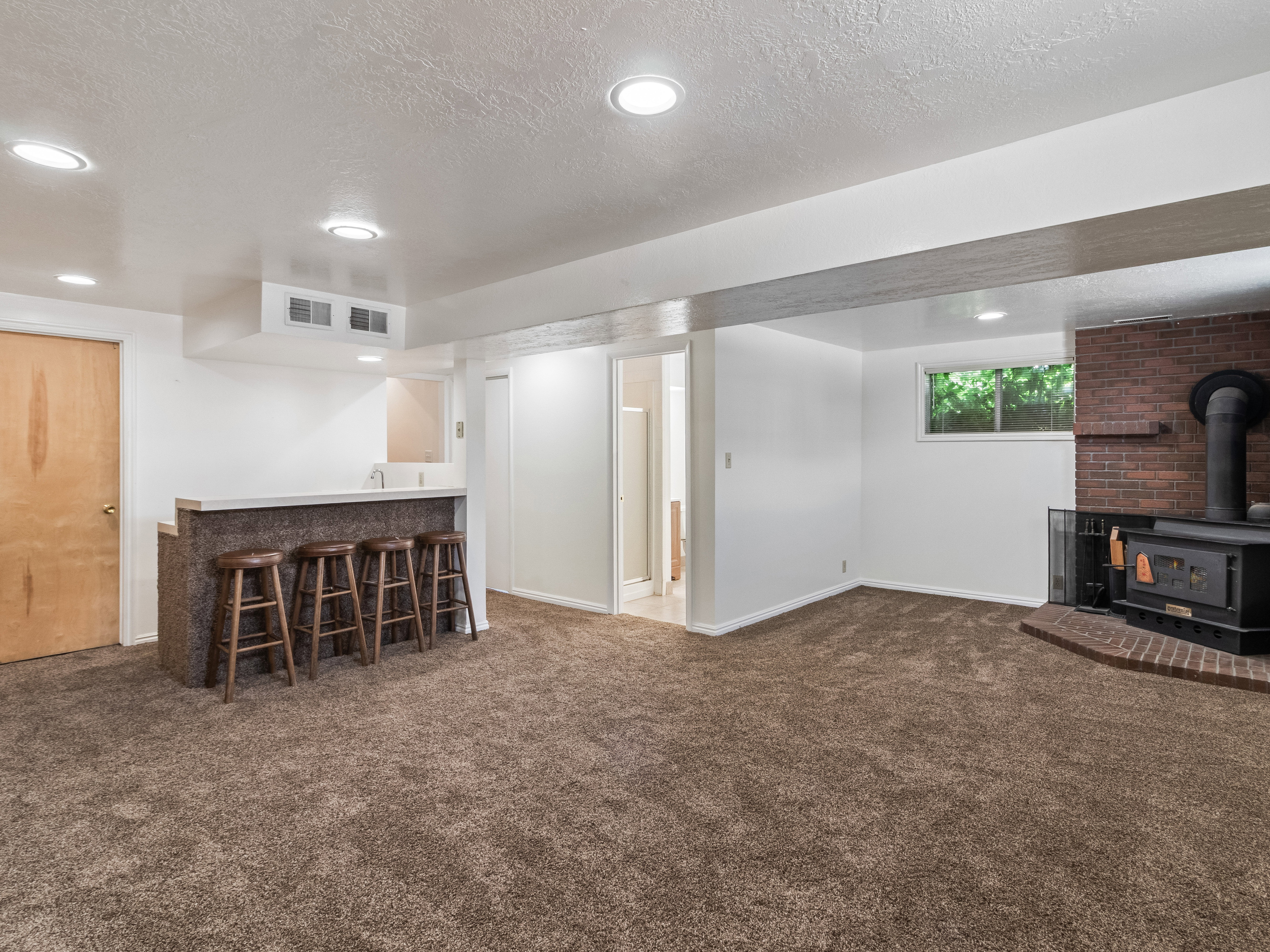 Spacious room featuring a built-in wet bar with a countertop and sink, a brick fireplace with a wood-burning stove, brown carpet flooring, and recessed ceiling lighting