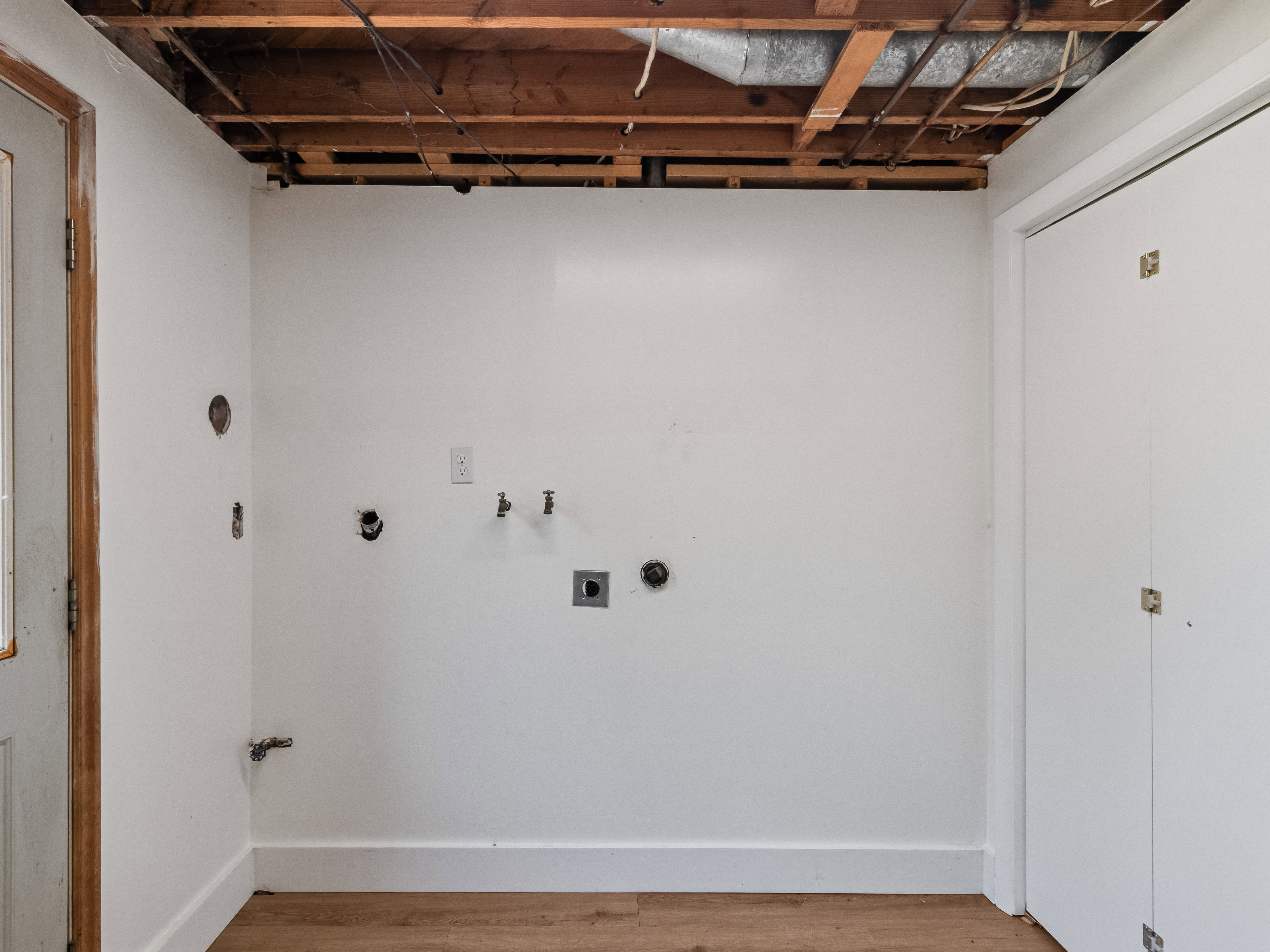 Laundry area featuring white walls, wood-finish flooring, exposed ceiling joists, and bifold closet doors
