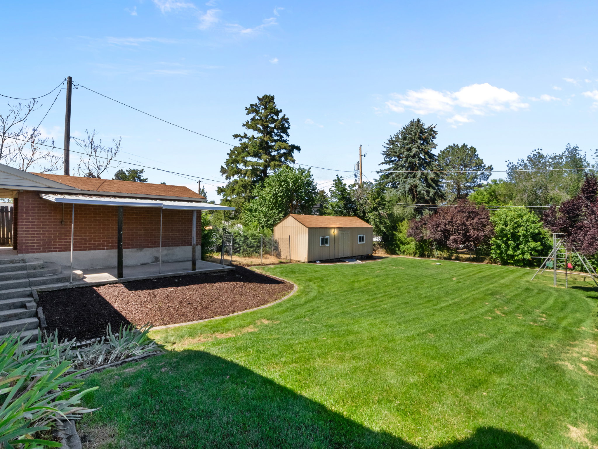 Spacious backyard featuring a covered patio with brick siding, a manicured lawn, a detached storage shed, a mulched planting area, and mature perimeter trees