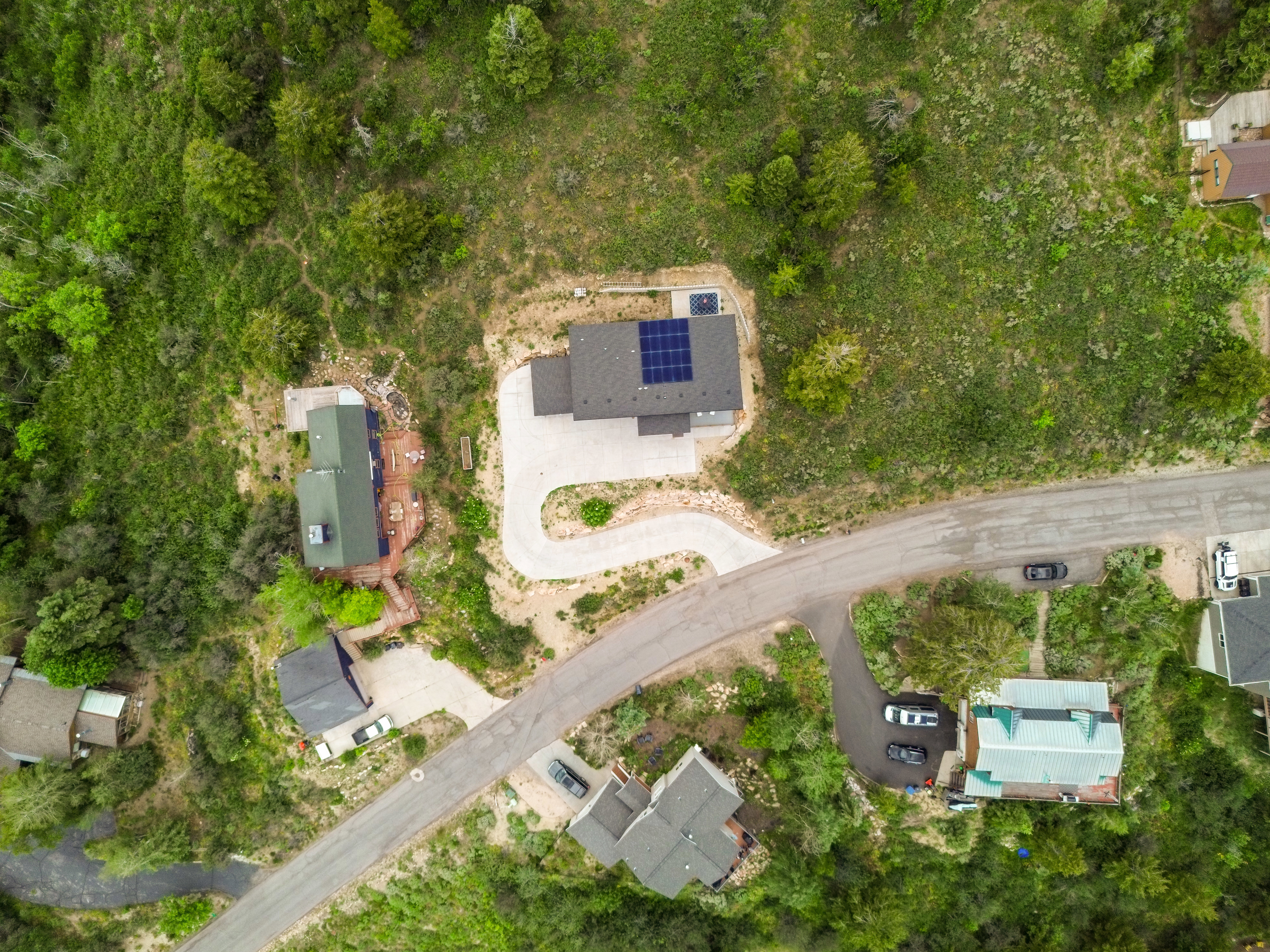 Aerial depiction of a property featuring a curved asphalt driveway, solar panels on the roof, a dark gray roofline, and surrounding natural foliage