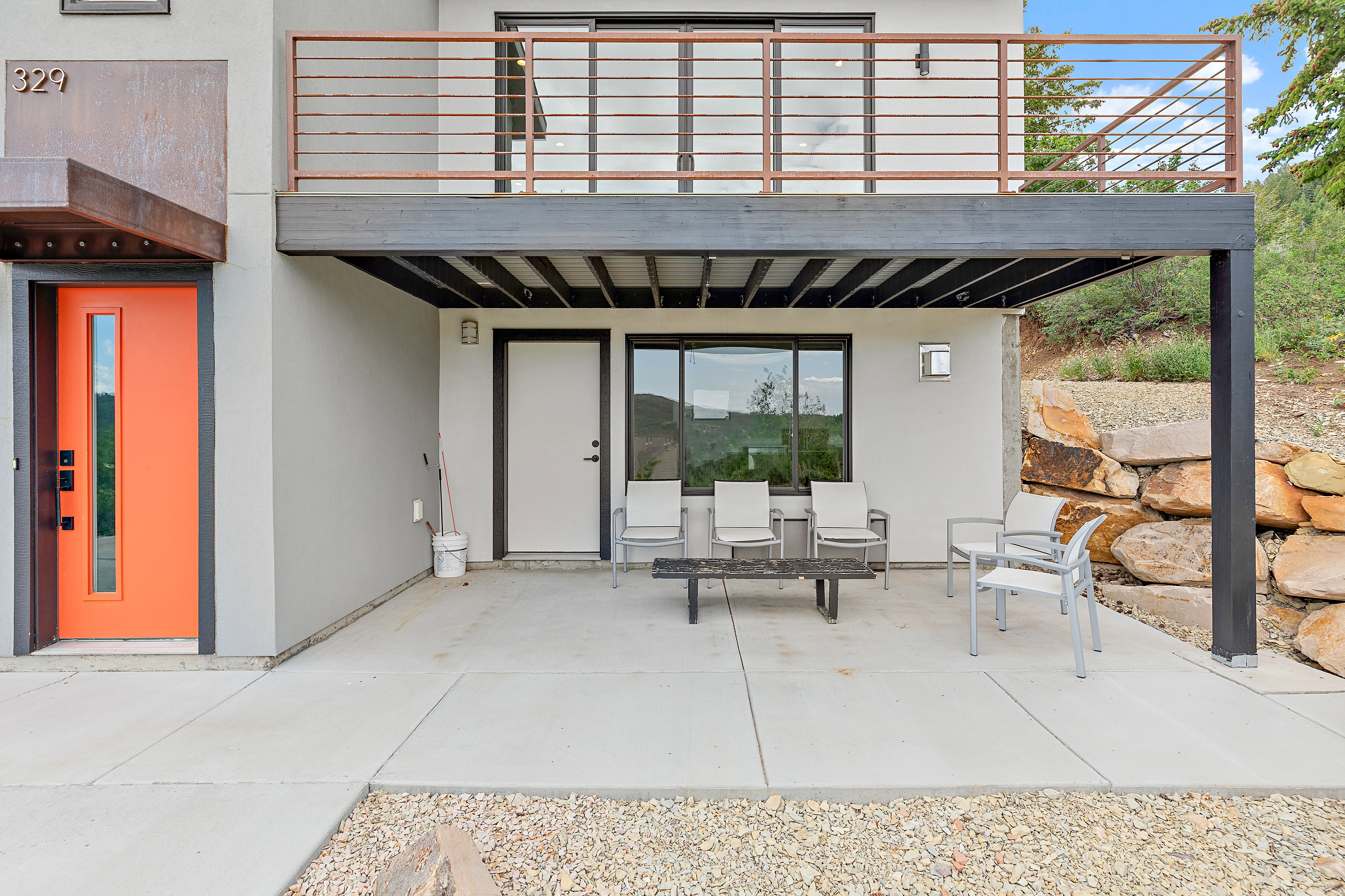 Modern exterior showcasing an orange entry door with a vertical glass insert, a steel balcony with horizontal railing, and a ground-level concrete patio