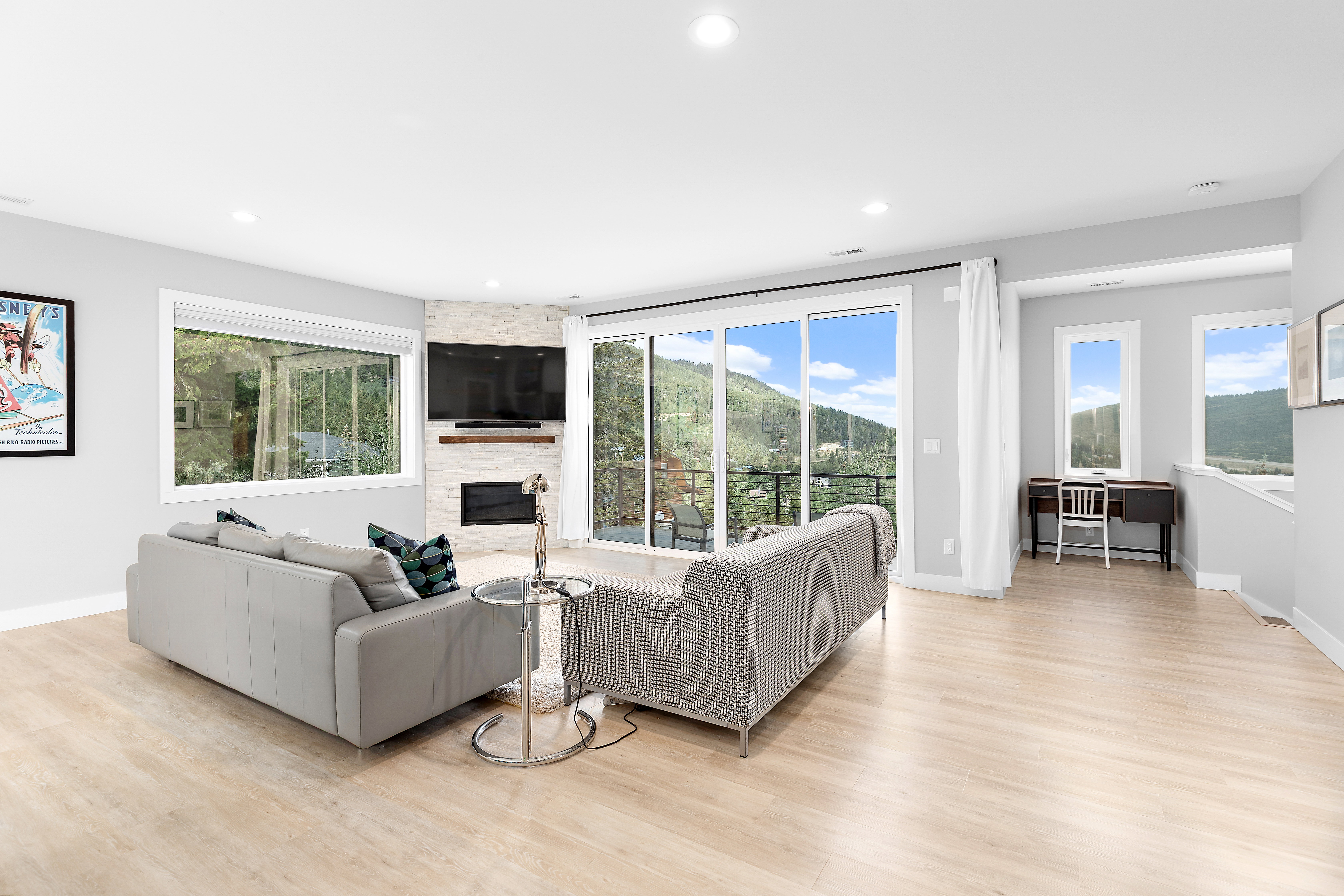Spacious living area featuring light wood-finish flooring, a corner fireplace with a stacked stone facade and wood mantel, large sliding glass doors opening to a balcony, and abundant natural light from multiple windows
