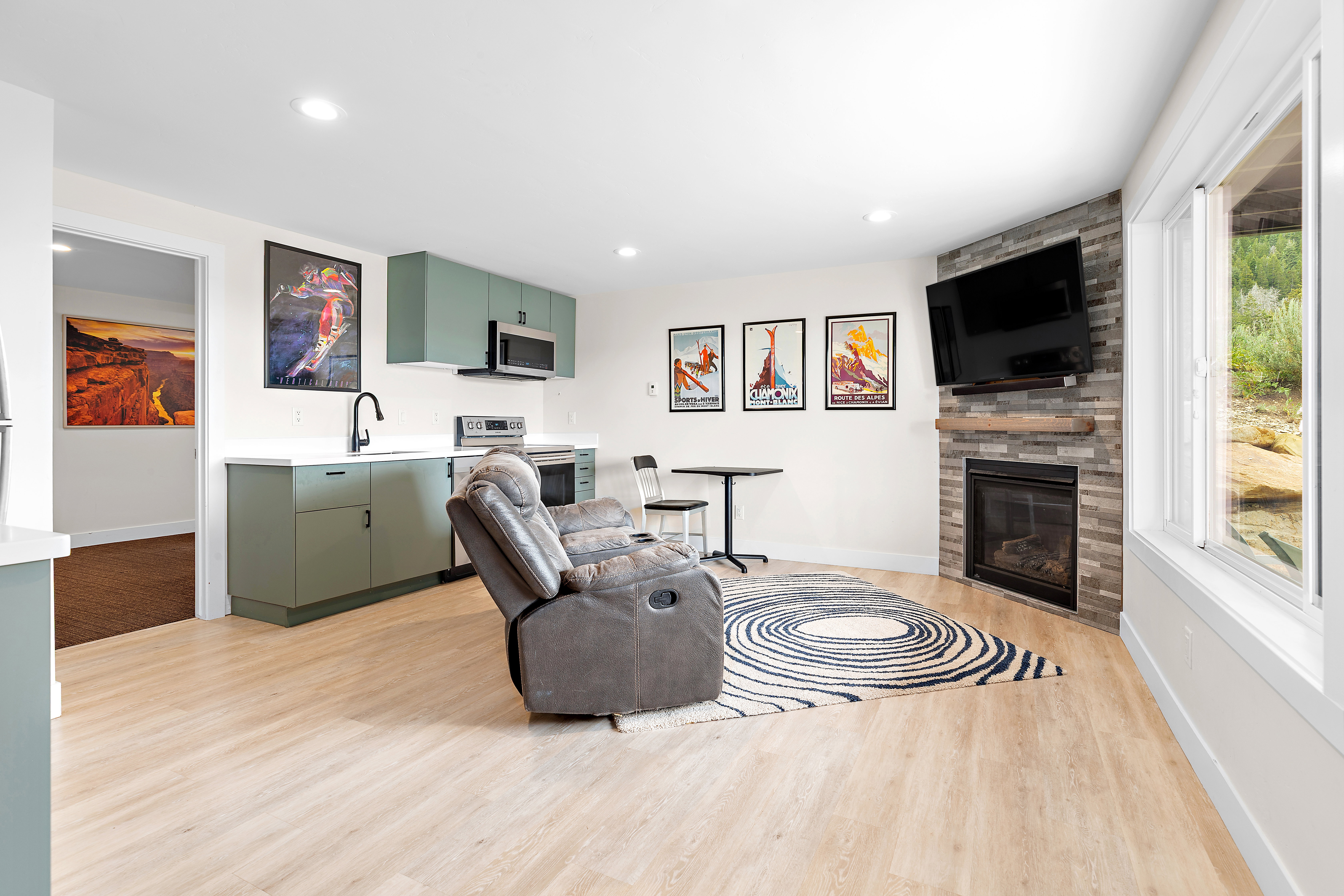 Open-concept living space featuring wood-finish flooring, a contemporary fireplace with a stone-tile surround, and a compact kitchen with green cabinetry and a white countertop