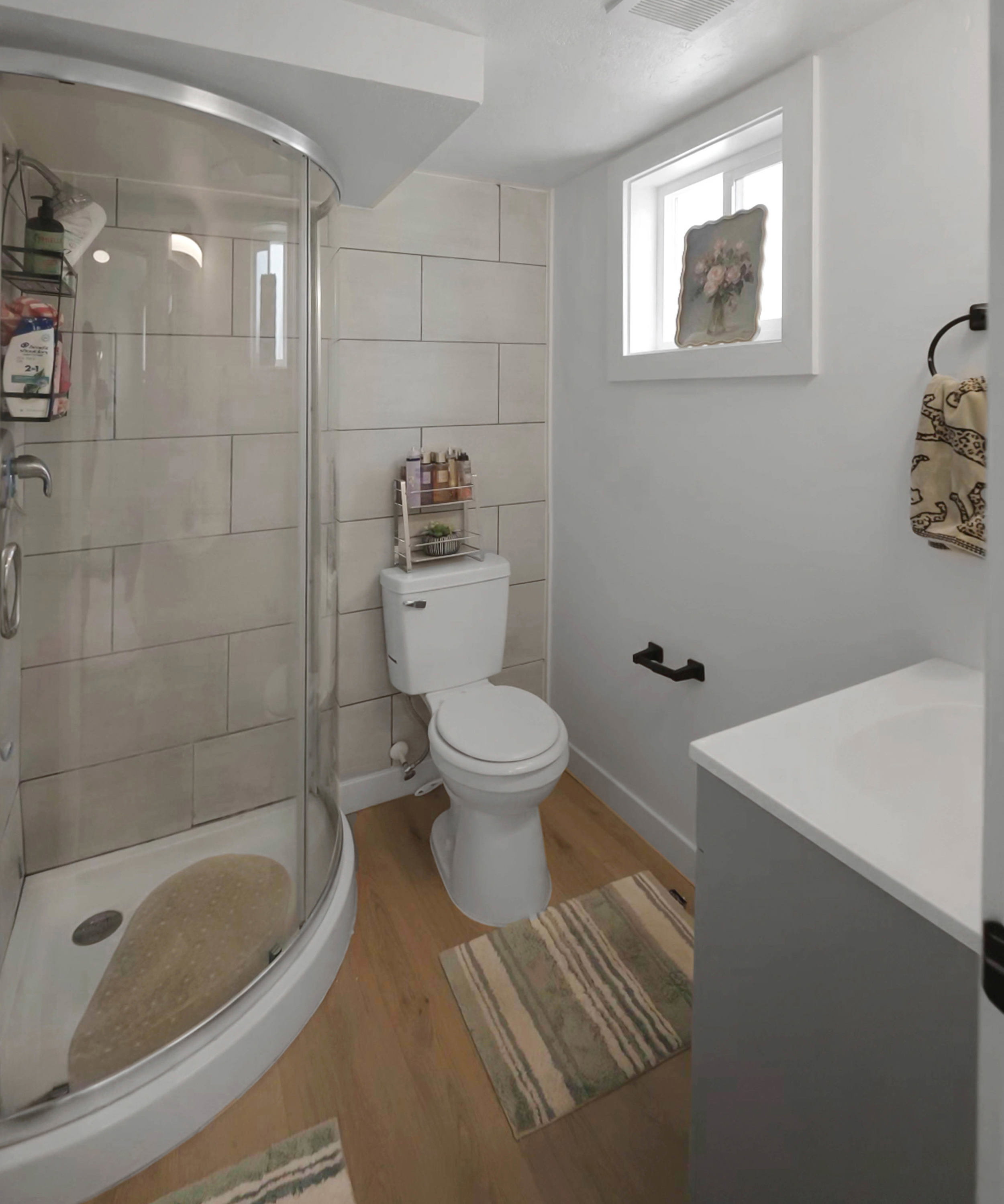 Three-quarter bathroom featuring a corner shower with glass enclosure and tiled walls, a white toilet, a window with white trim, a single-sink vanity with a light-colored countertop, and wood-finish flooring