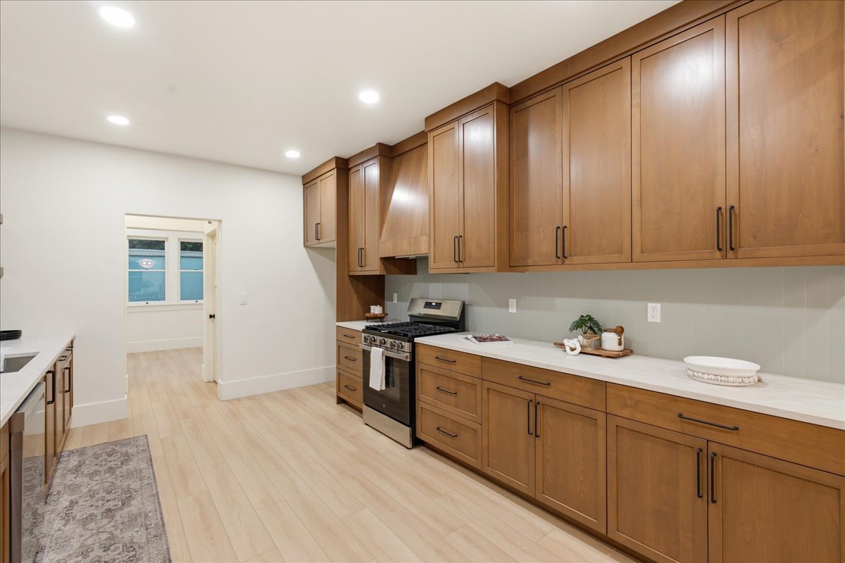 Contemporary kitchen featuring light wood-finish flooring, extensive wood cabinetry with dark hardware, light countertops, a stainless steel range with a matching overhead vent hood, and recessed ceiling lighting