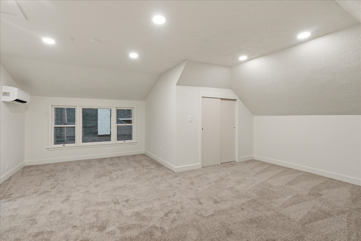 Spacious attic room featuring plush grey carpeting, recessed ceiling lighting, a triple window unit with white trim, a wall-mounted air conditioning unit, and built-in sliding closet doors
