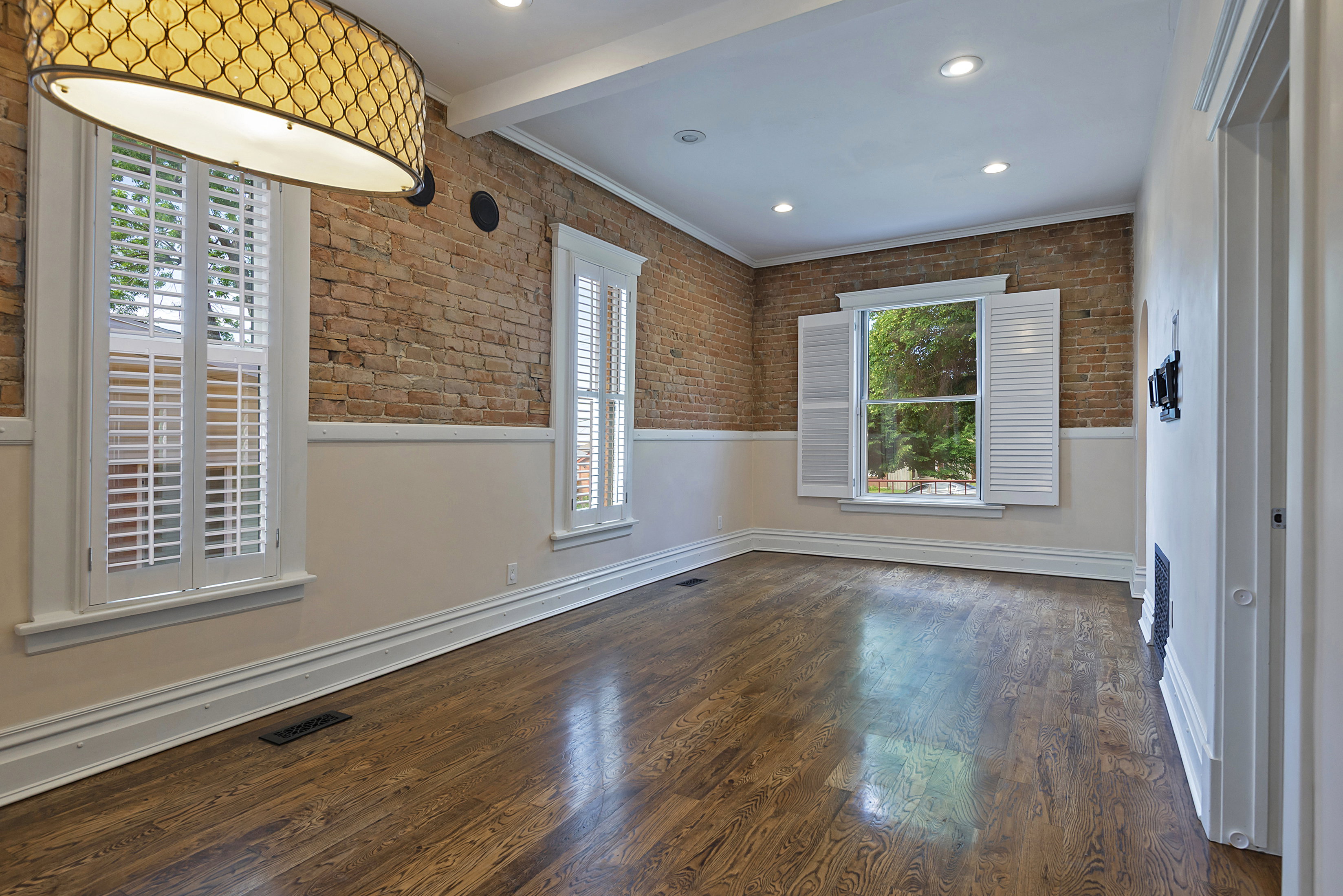 Room featuring exposed brick walls, wood-finish flooring, white plantation shutters, decorative ceiling light fixture, and recessed lighting
