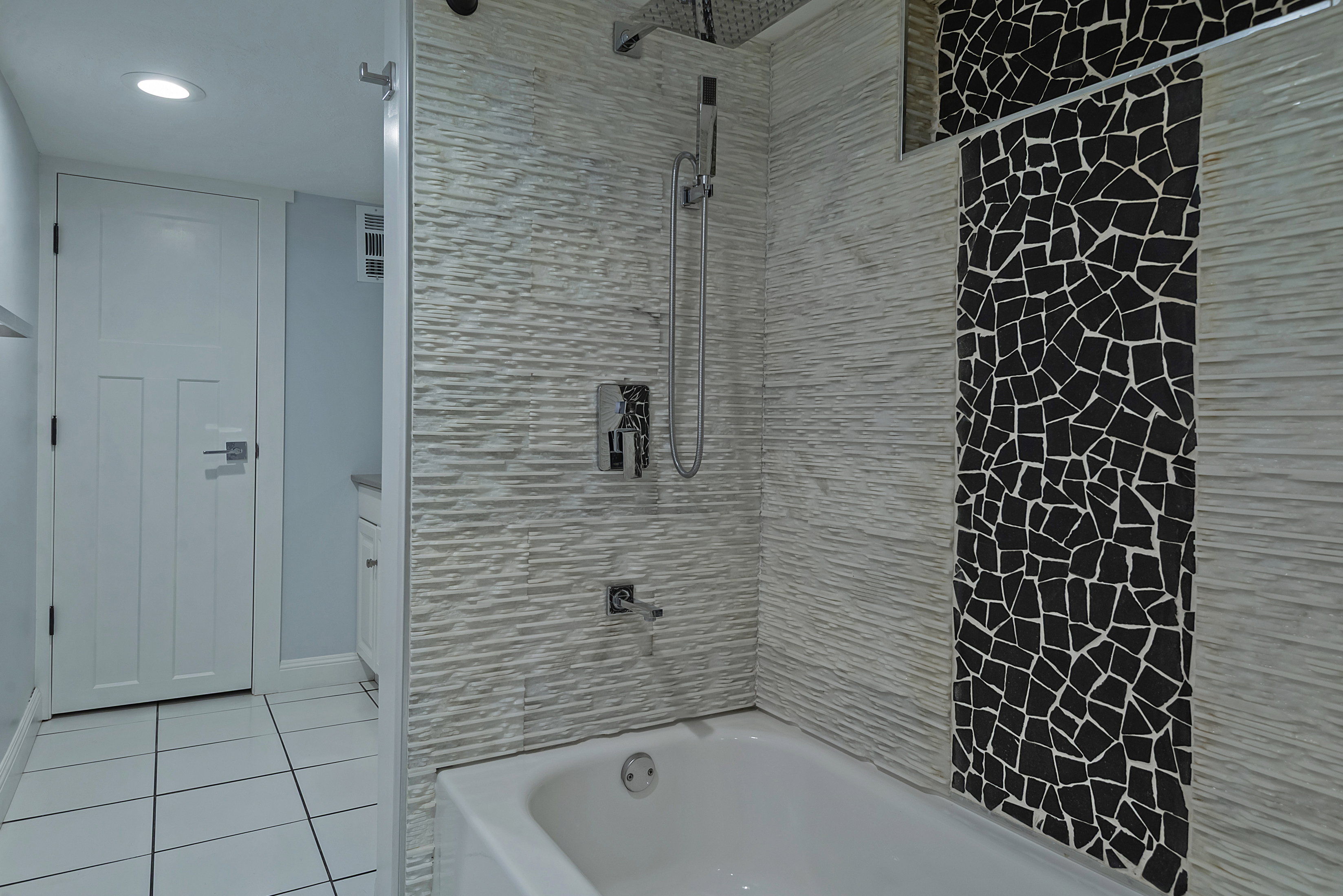 Bathtub with a textured white tile surround, a black mosaic tile accent strip, and a chrome shower fixture