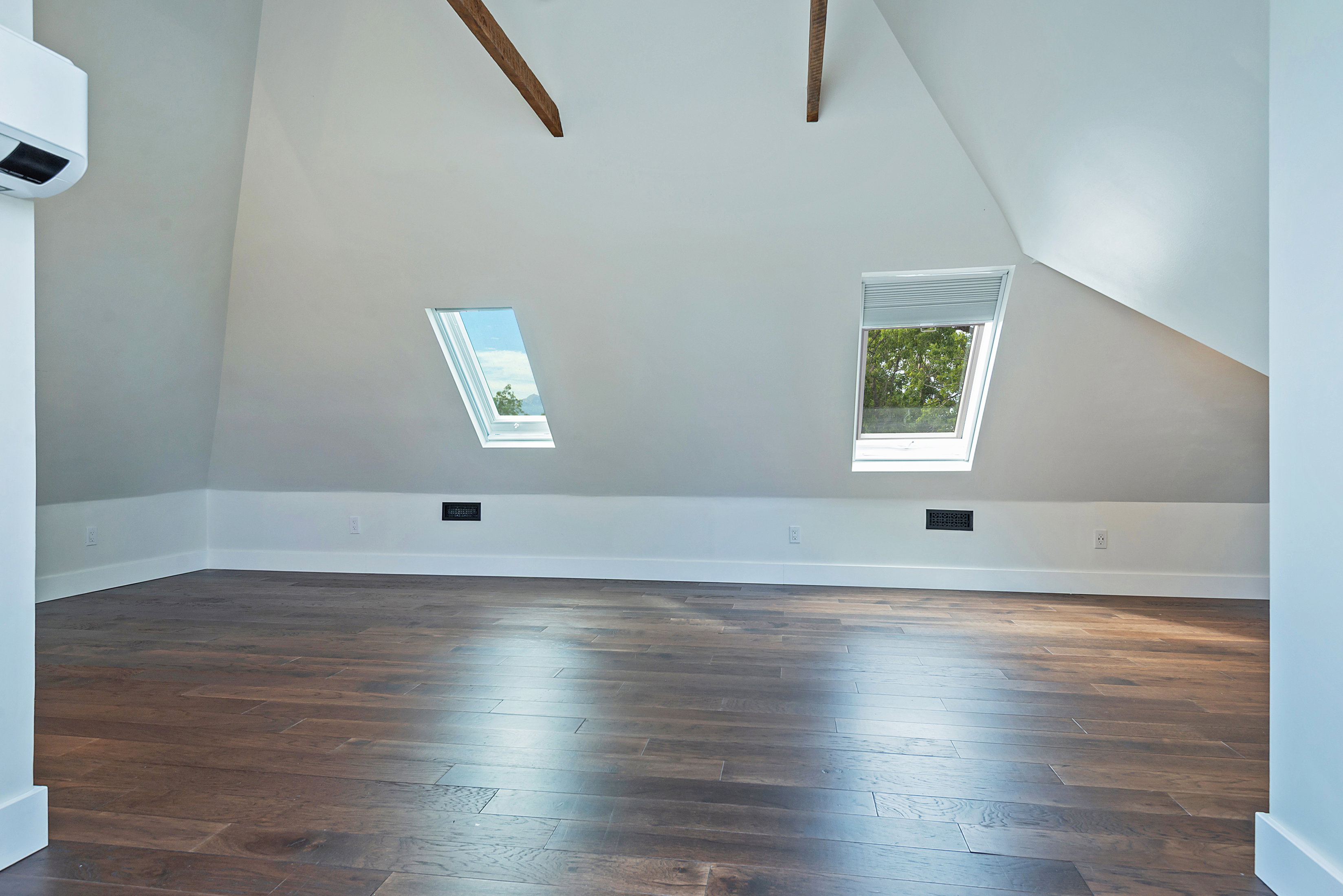 Spacious room featuring rich wood-finish flooring, two skylight windows, exposed ceiling beams, and a wall-mounted air conditioning unit