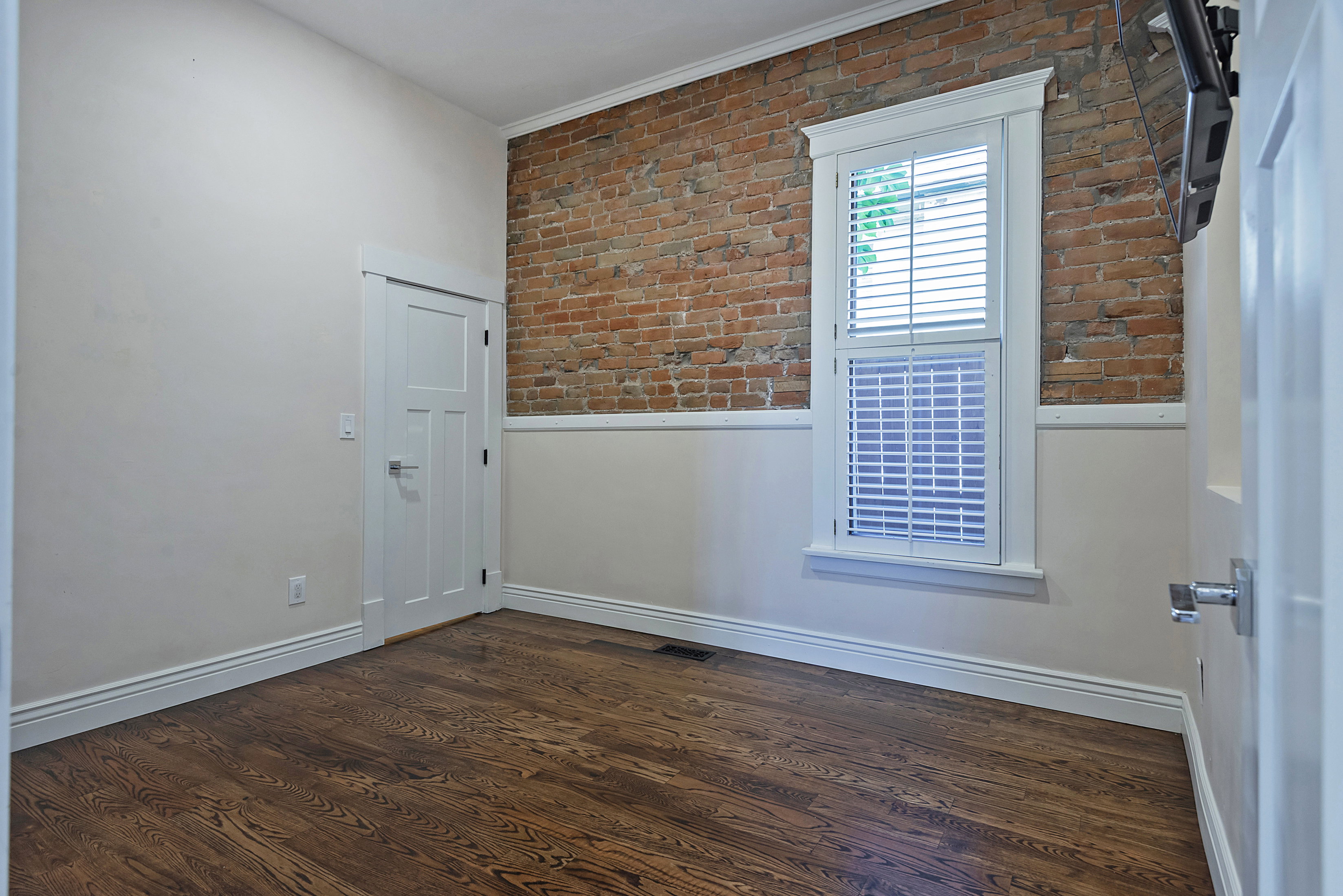 Exposed brick accent wall with a white trim window featuring louvered shutters, wood-finish flooring, and white baseboards