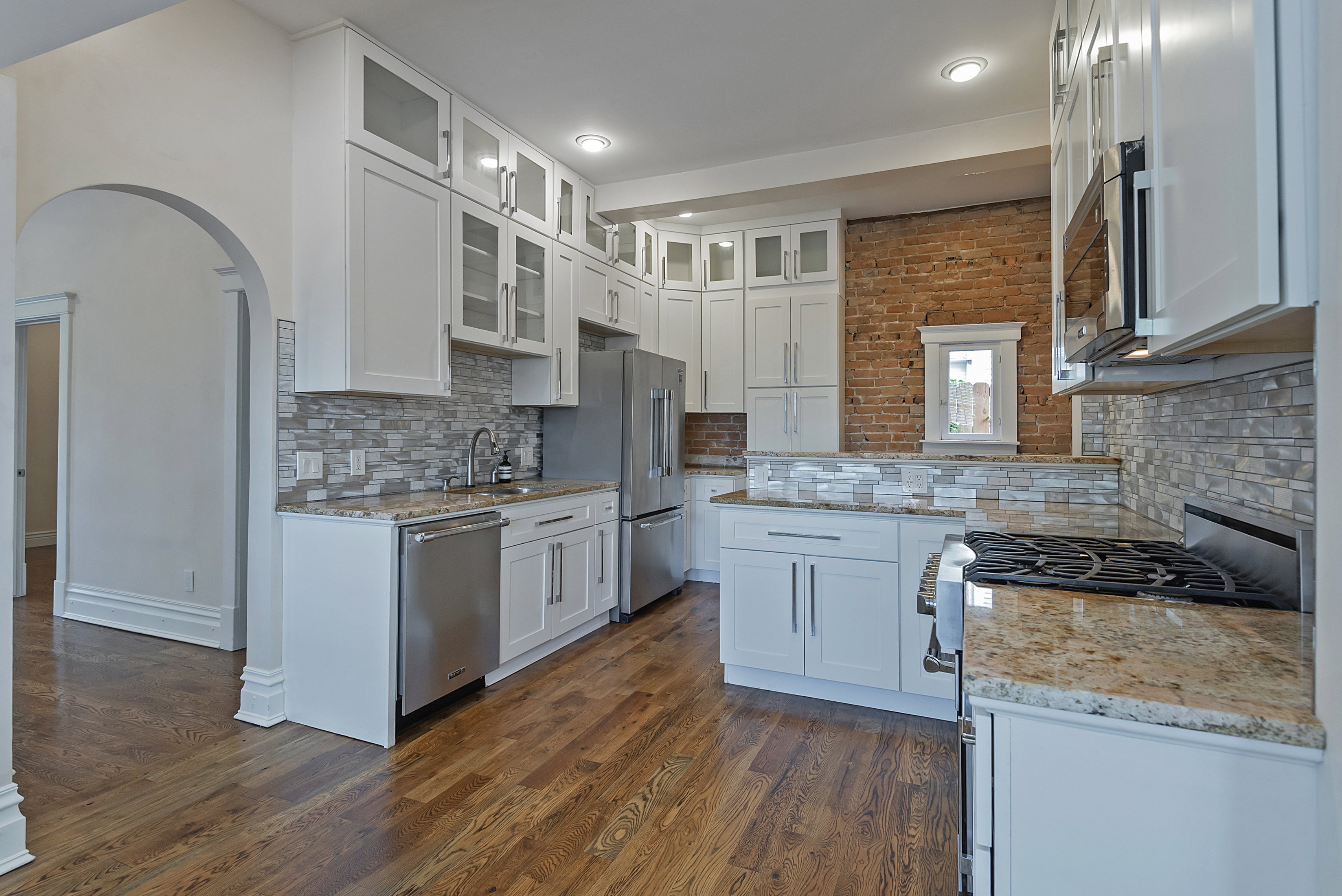 Open-concept kitchen featuring wood-finish flooring, white cabinetry, stainless steel appliances, stone countertops, and a full-height tile backsplash