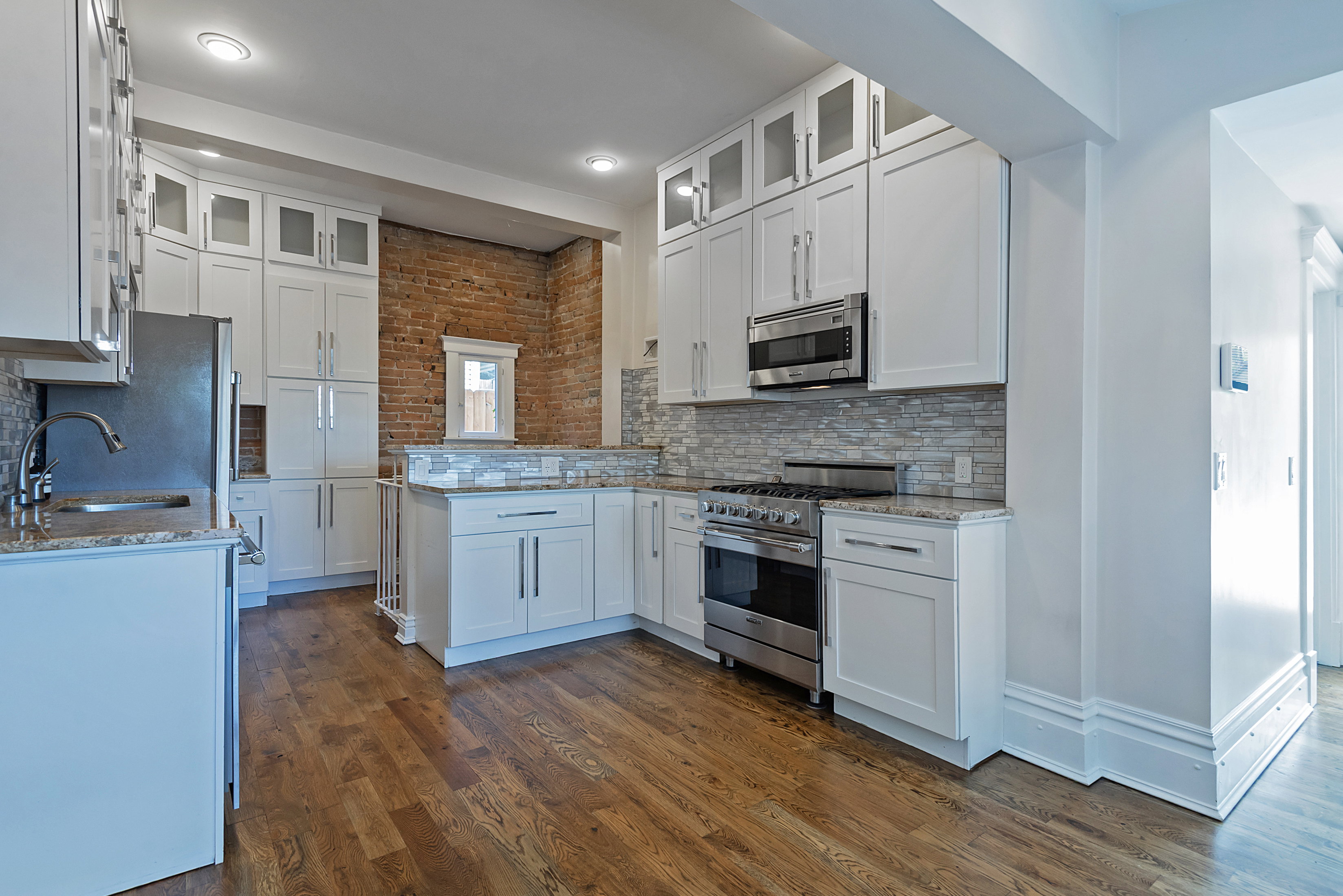 Kitchen featuring white cabinetry, stainless steel appliances, granite countertops, wood-finish flooring, and an exposed brick accent wall