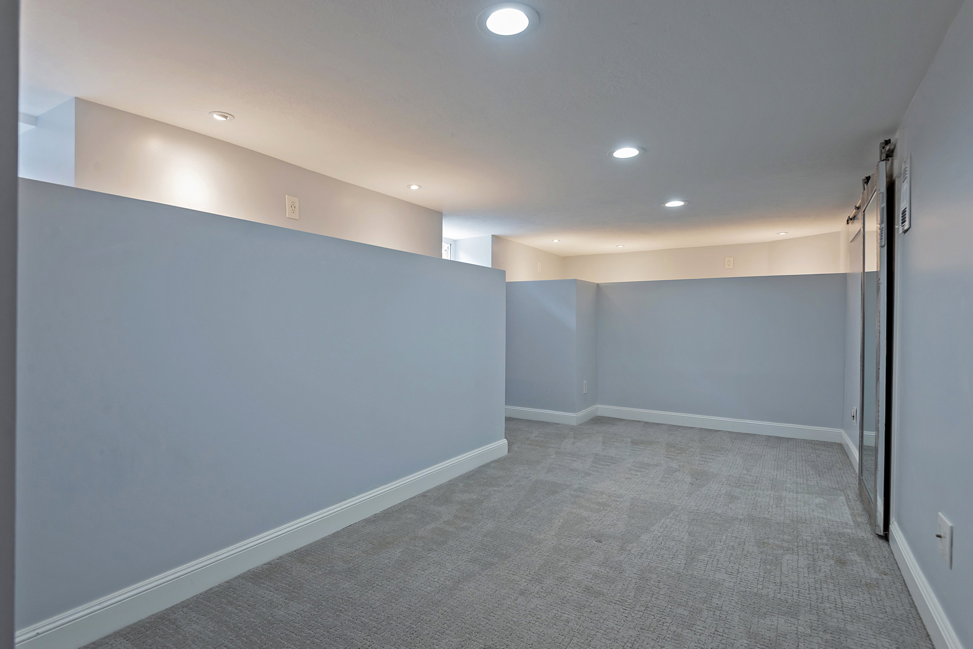 Spacious room featuring light gray walls, recessed lighting, gray patterned carpeting, white baseboards, and a barn door with a mirrored panel