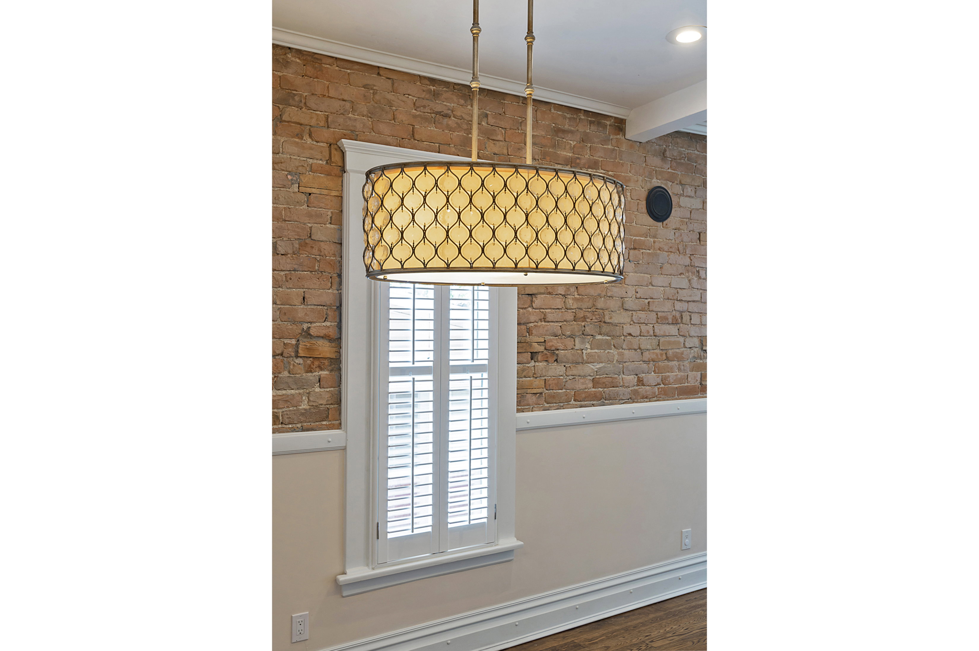 Exposed brick wall featuring a window with white plantation shutters, complemented by a decorative drum pendant light fixture and recessed ceiling lighting