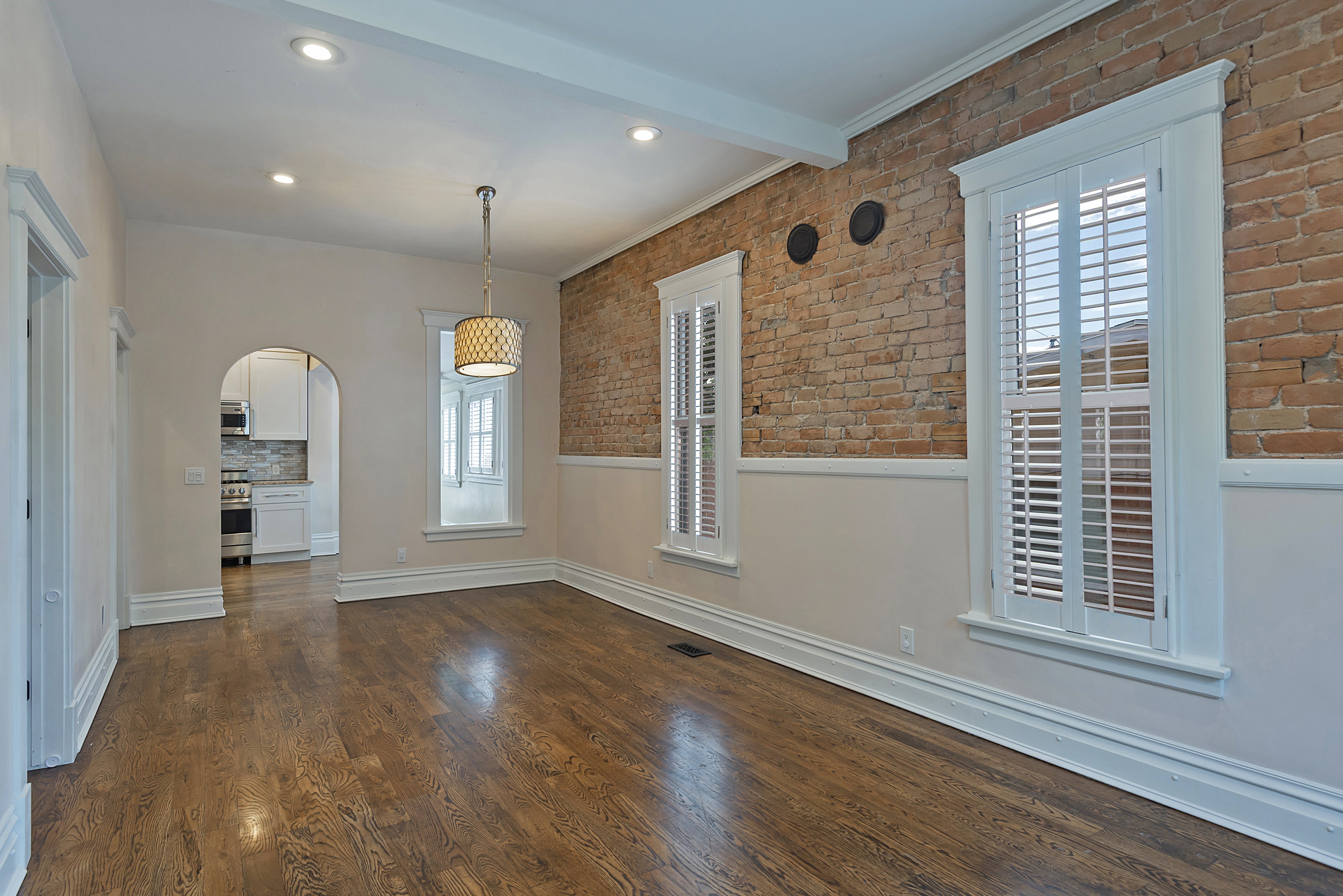 Spacious room featuring exposed brick, wood-finish flooring, white plantation shutters, an arched doorway, and recessed lighting