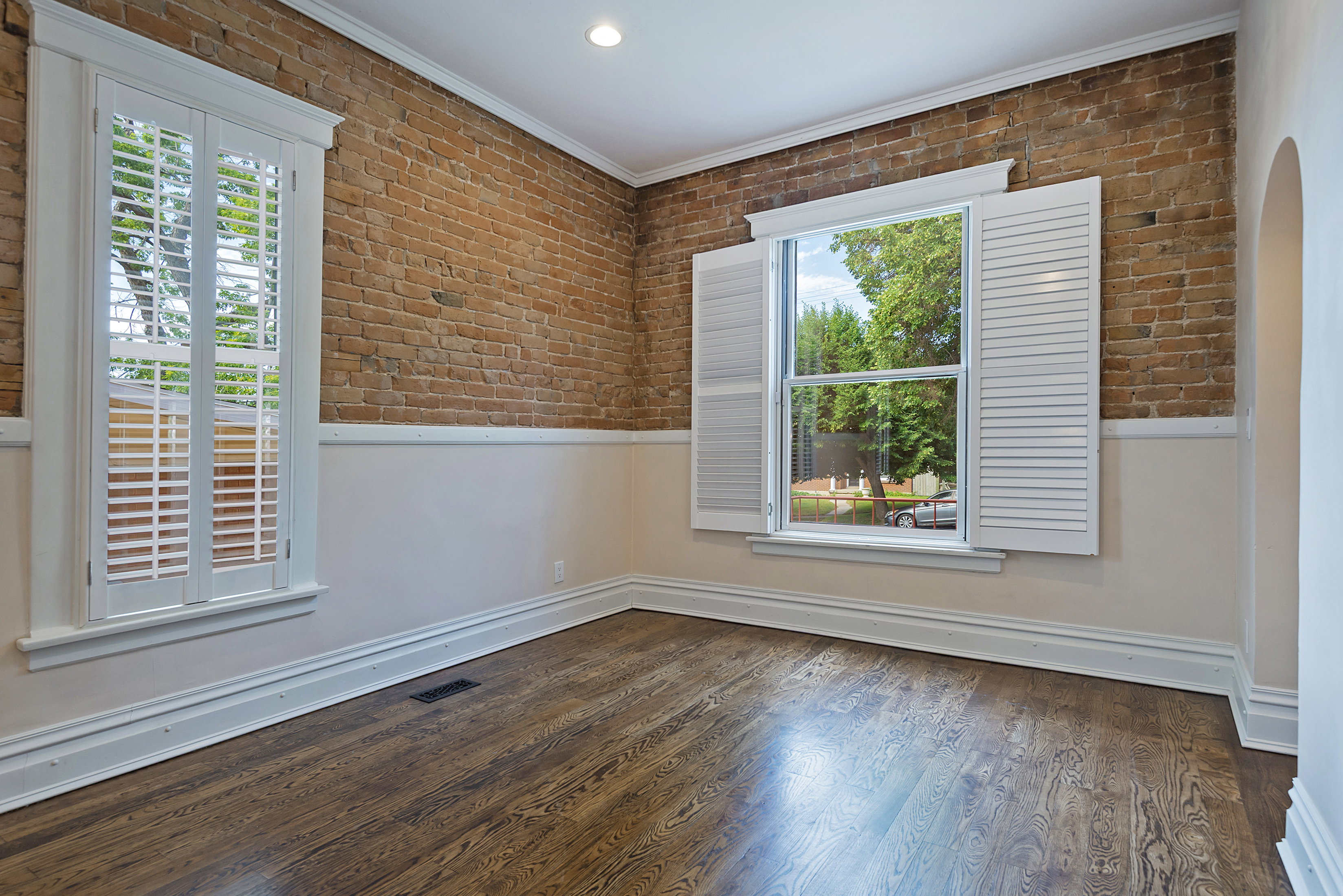 Exposed brick wall featuring a window with white louvered shutters, crown molding, wood-finish flooring, and a wainscoting detail