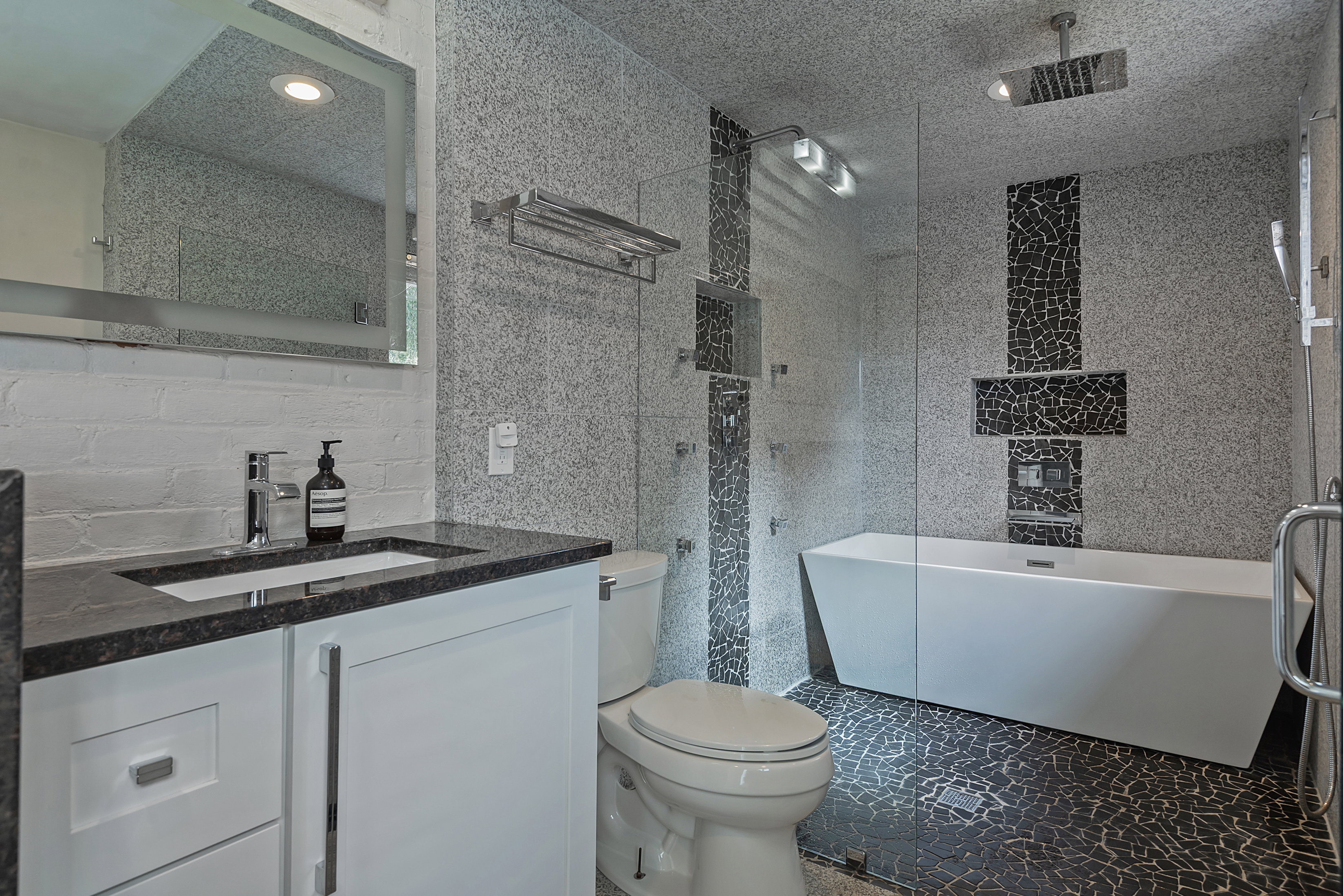 Modern bathroom featuring a freestanding tub, glass-enclosed shower, white vanity with a dark countertop, white brick-patterned wall, and textured gray and black tile
