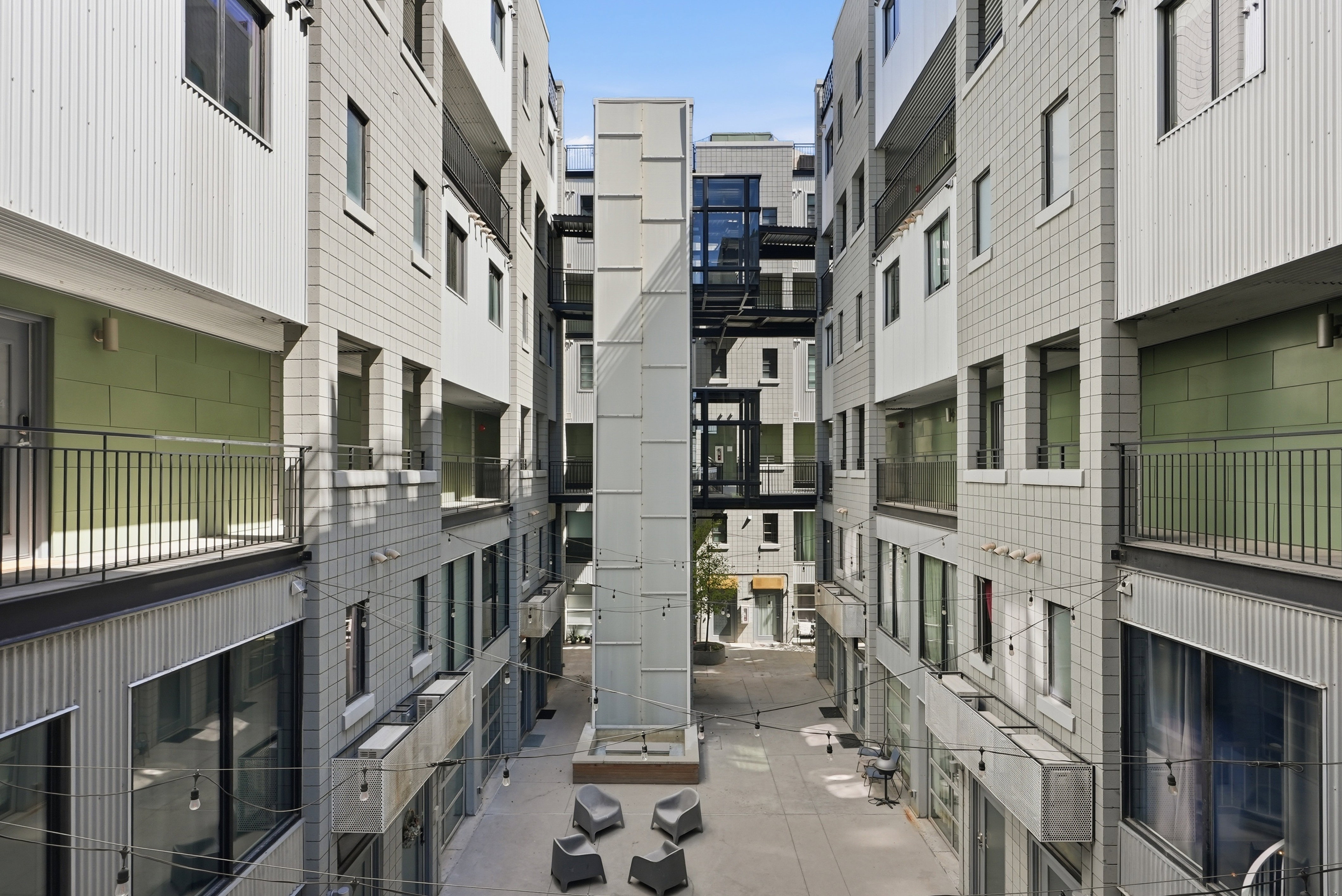 Modern multi-level building exterior featuring corrugated metal and square-paneled siding, multiple balconies with dark railings, a central glass-enclosed elevator shaft, and an open-air courtyard with seating