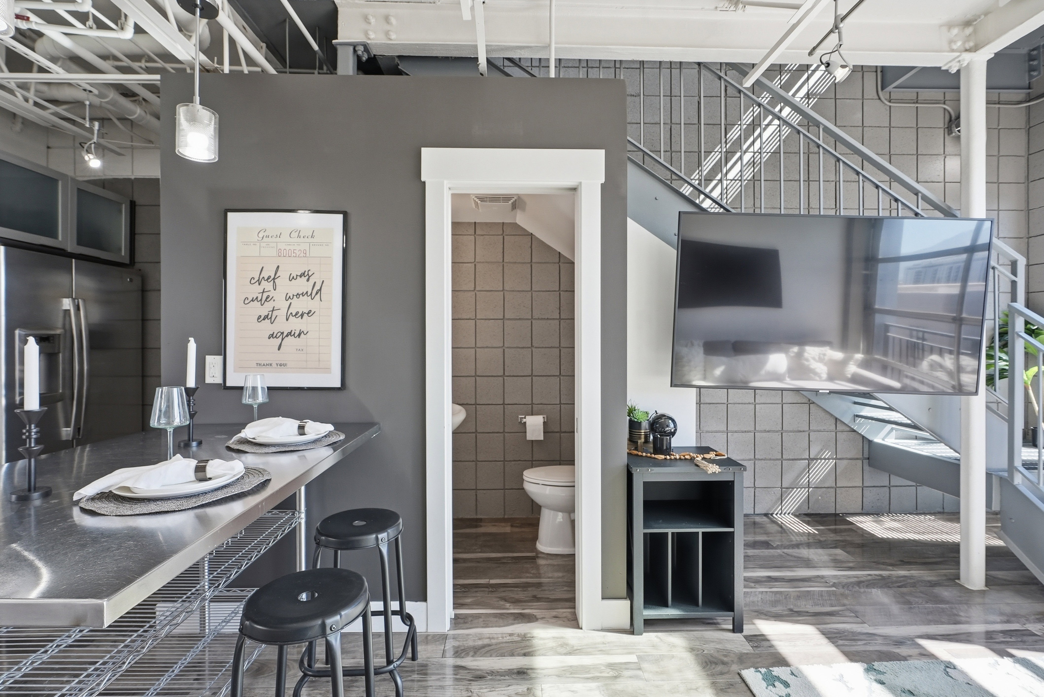 Open-concept living space featuring a stainless steel kitchen island with seating, exposed industrial-style ceiling beams, a contemporary open staircase, and wood-finish flooring