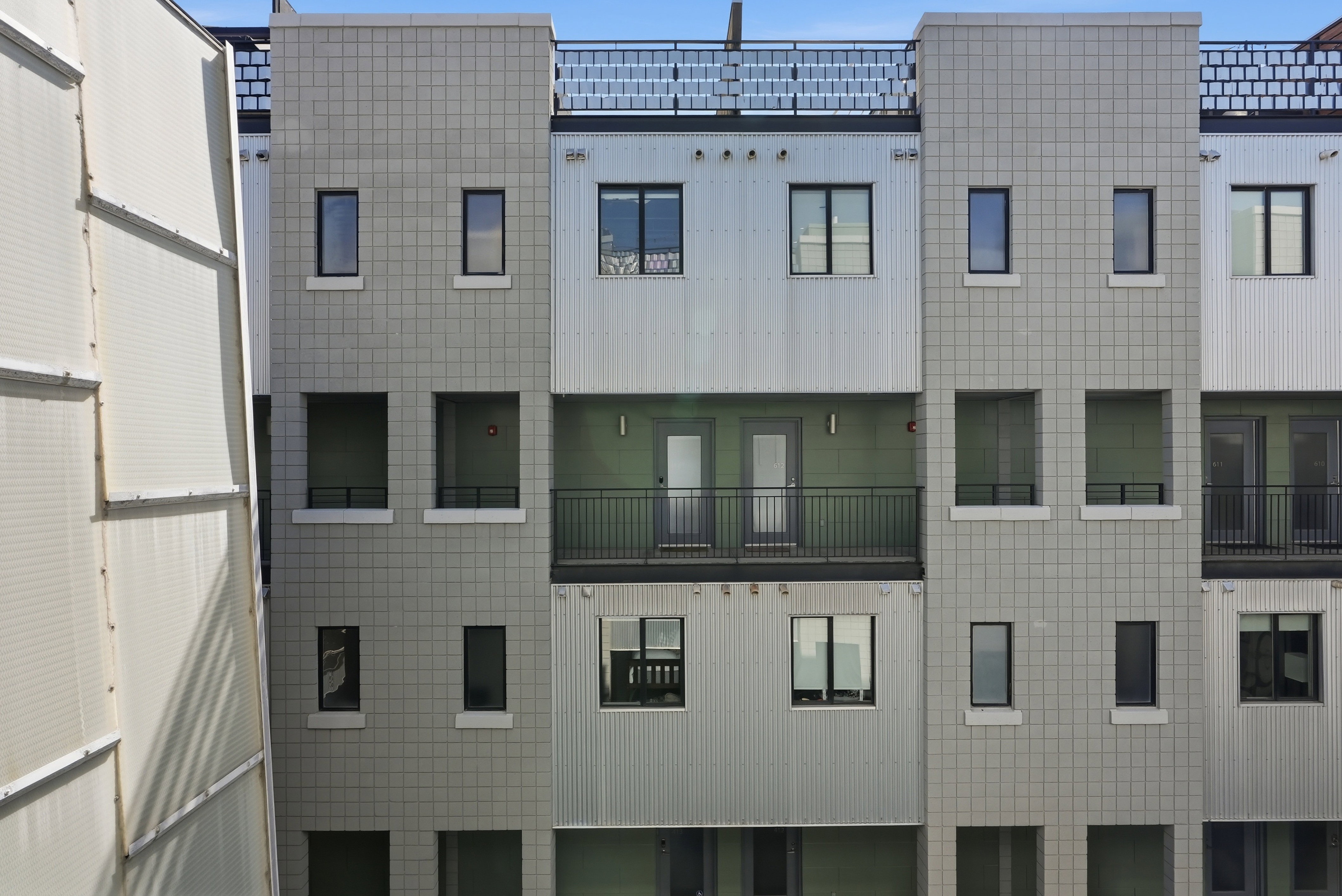 Multi-level contemporary facade featuring a central balcony with dark metal railings, contrasting smooth and corrugated paneling, and multiple dark-framed windows