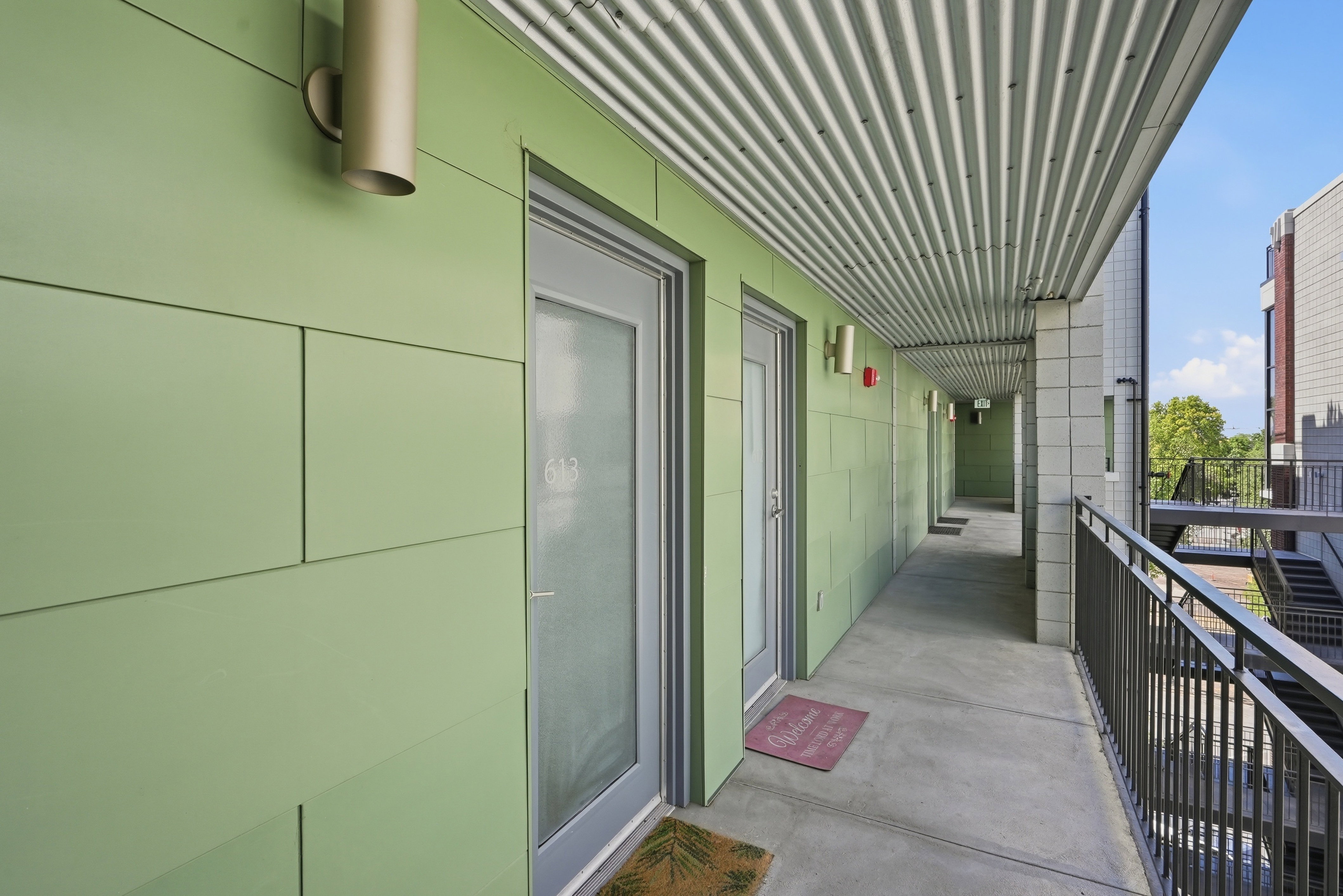 Open-air corridor featuring green paneling, corrugated metal ceiling, concrete flooring, and metal railings