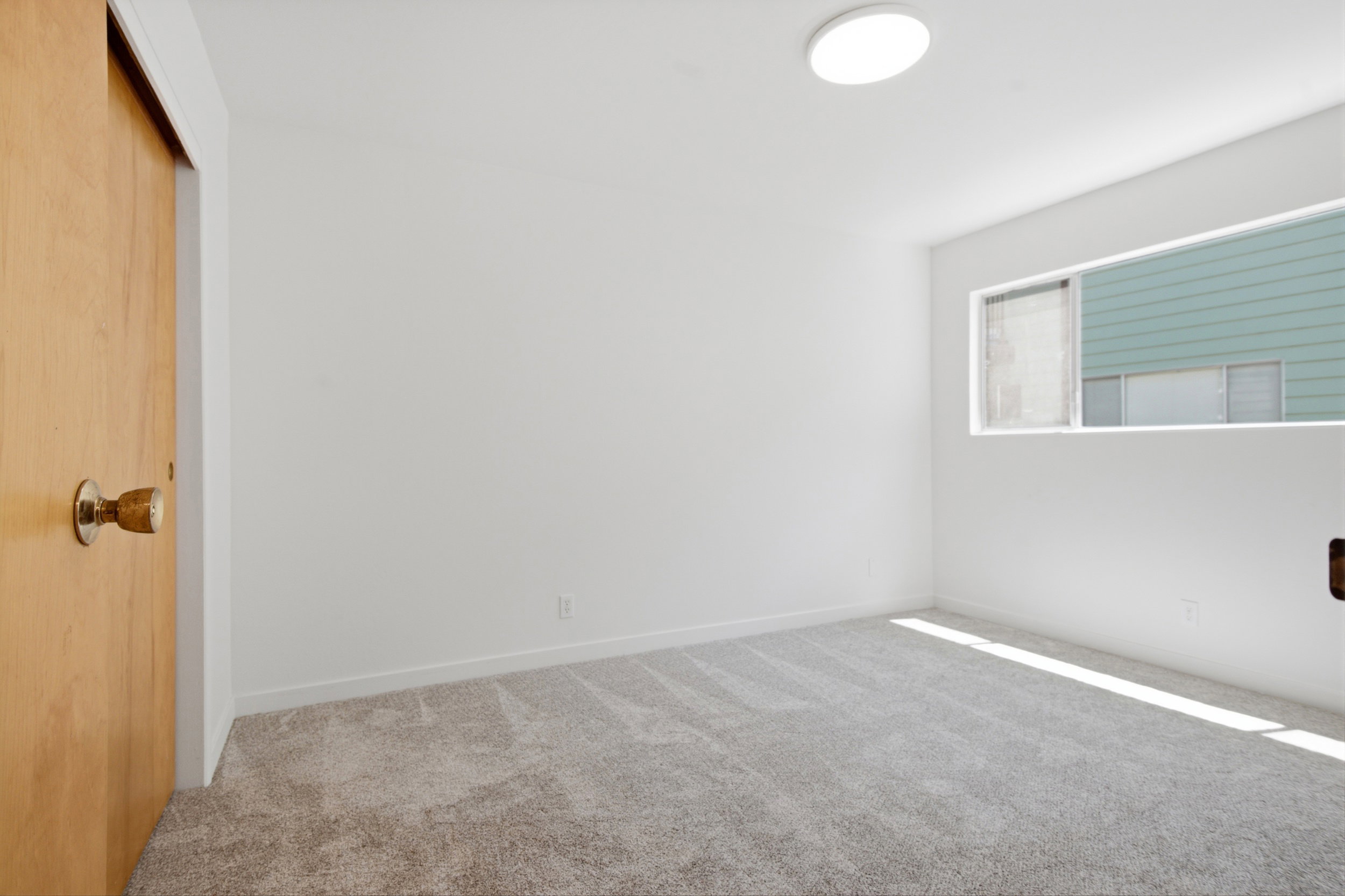 Spacious room featuring light-toned carpeting, white painted walls, a sliding wood-finish closet door, and a horizontal window providing natural illumination