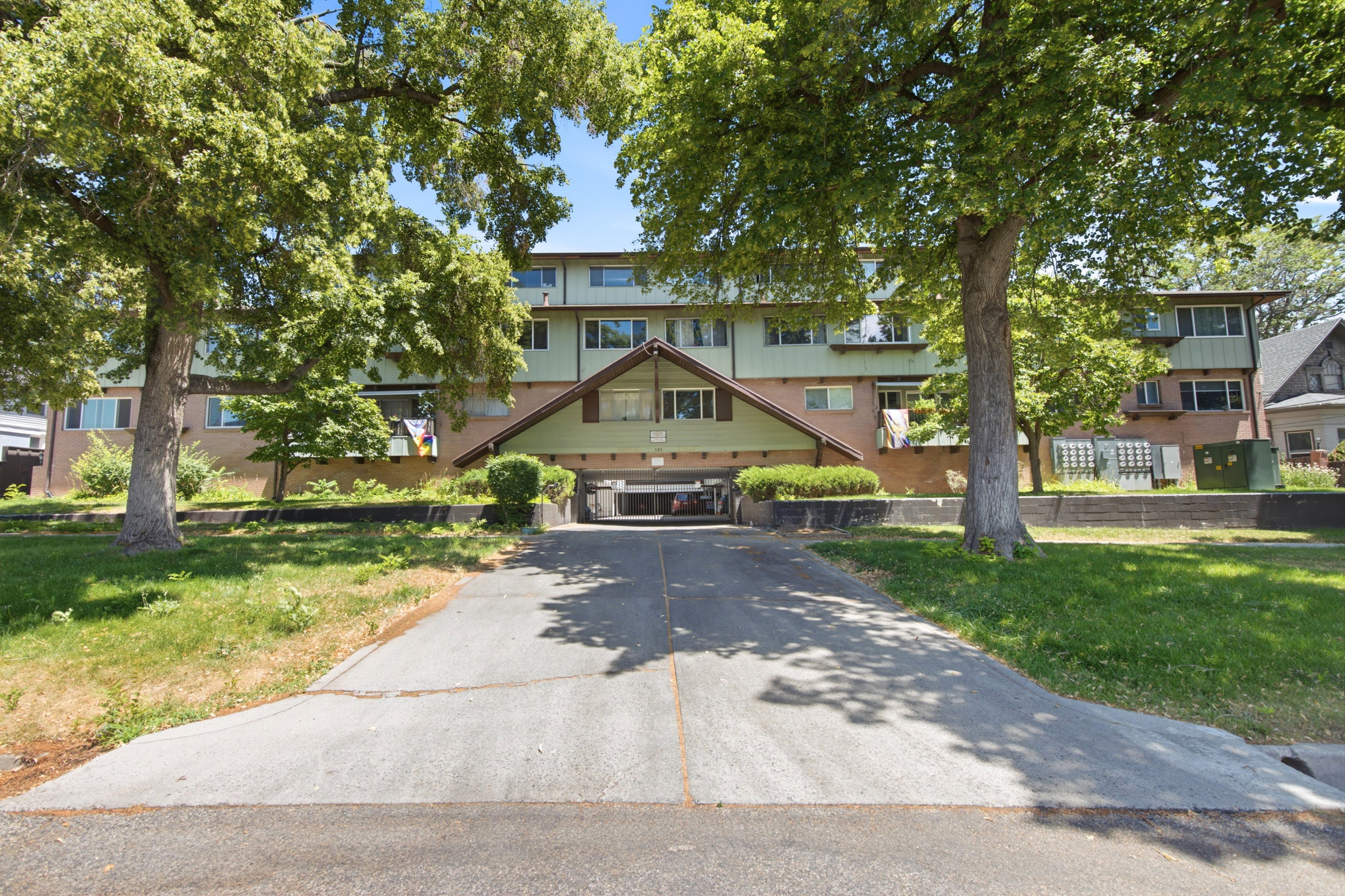 Multi-story building featuring brick and horizontal siding, mature trees, and a paved driveway leading to covered parking