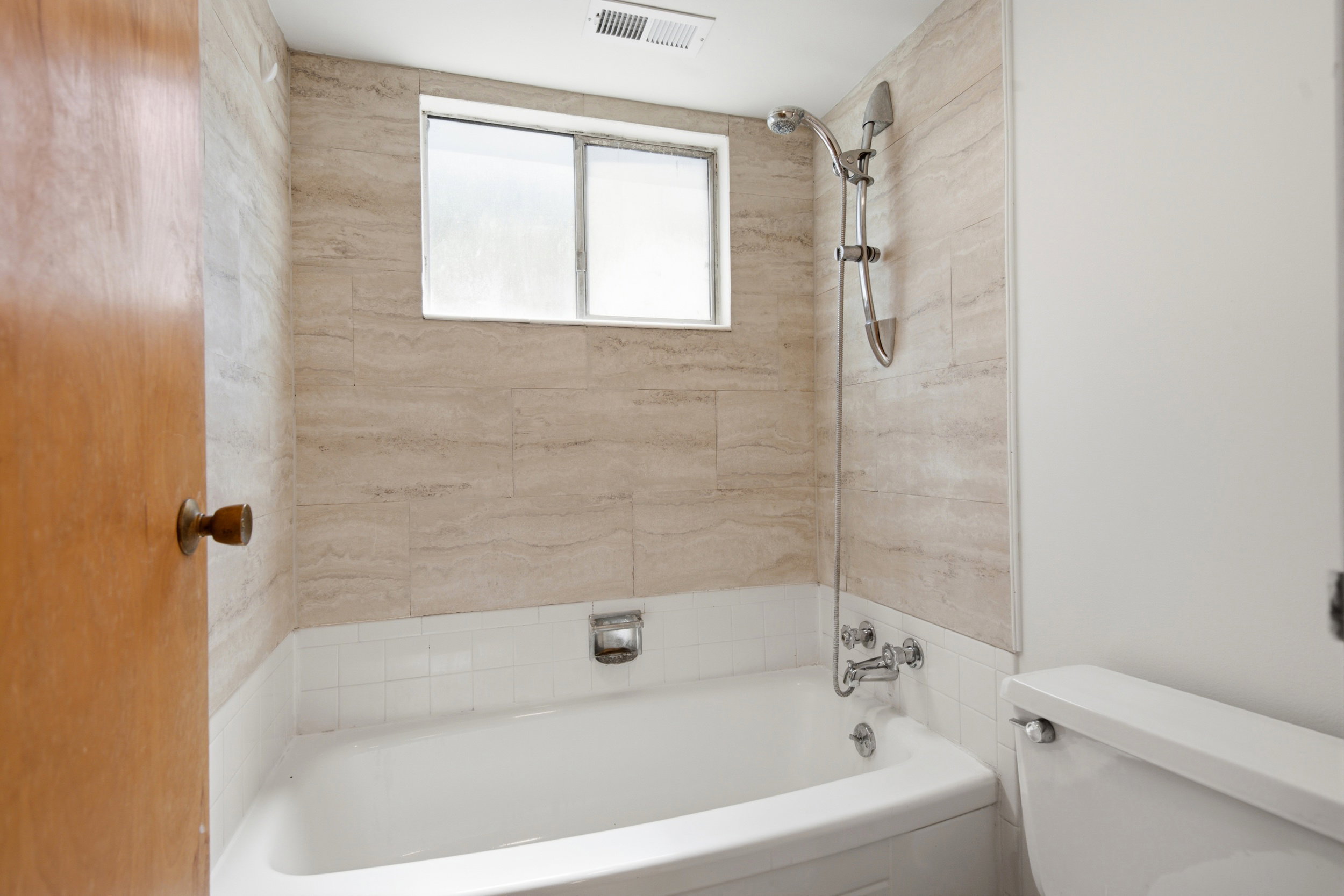 Bathtub and shower combination featuring neutral-toned rectangular wall tiling, a white tub, and a window with frosted glass