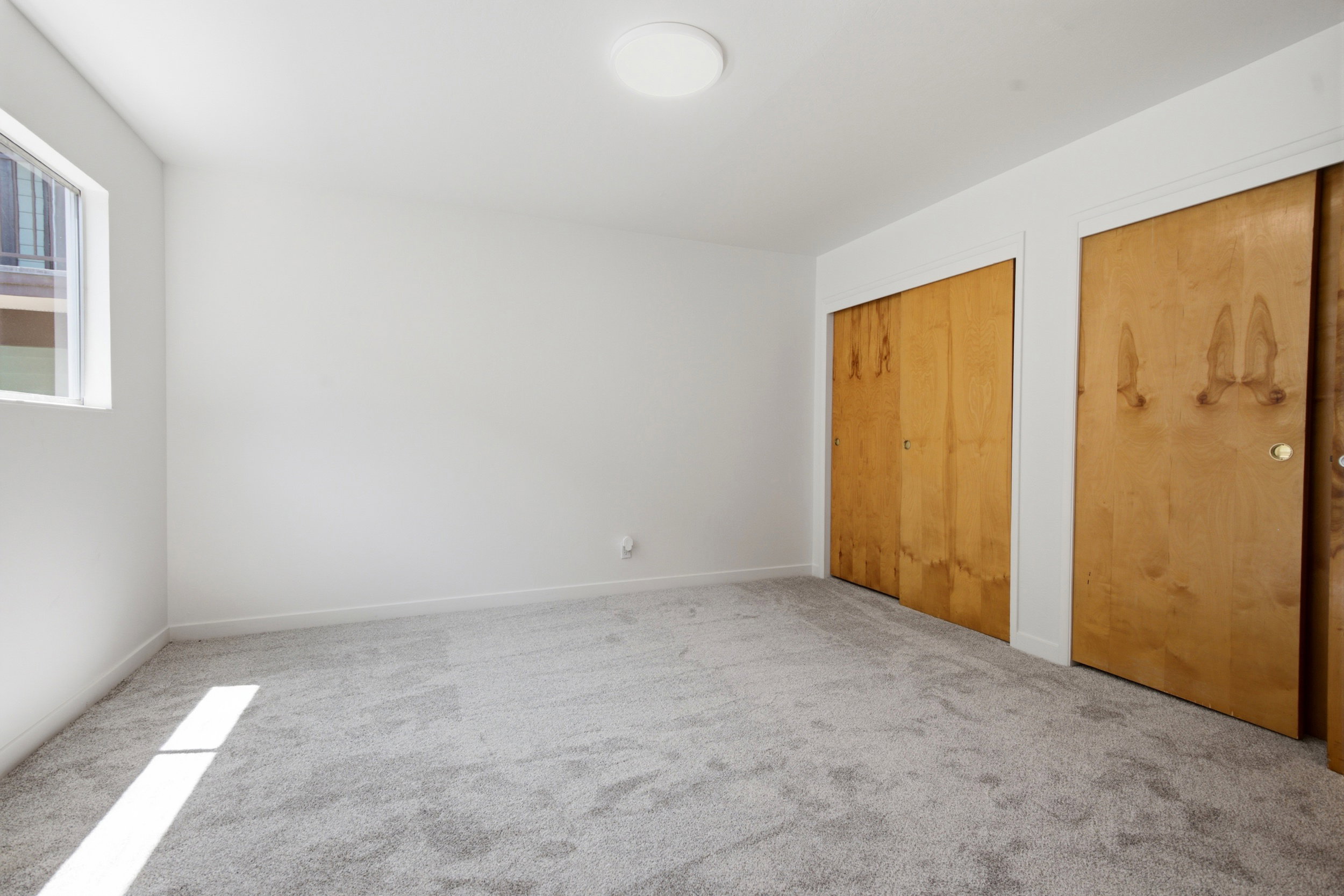 Light gray carpeted room featuring white walls, a single window, two wood-paneled sliding closet doors, and a flush-mount ceiling light fixture