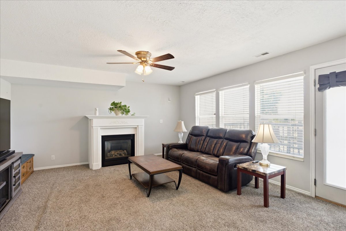 Living area featuring a white mantel fireplace, three large windows, a ceiling fan, light gray wall paint, and neutral carpet flooring