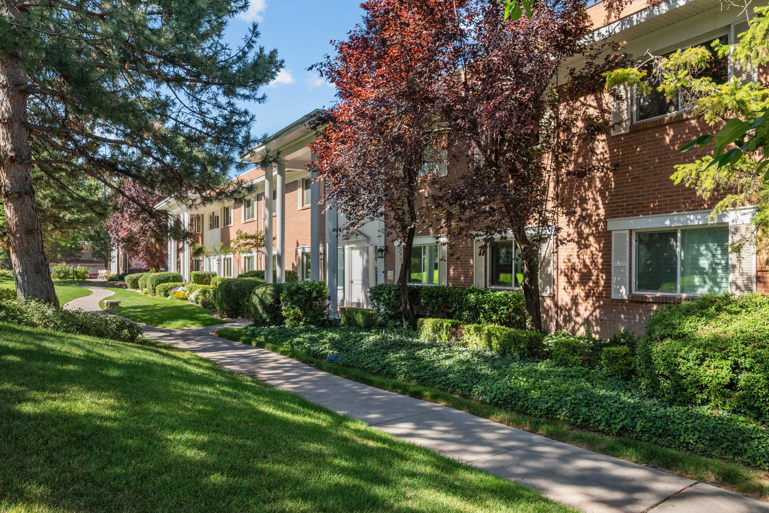 Brick exterior featuring white columns, white window shutters, mature trees, manicured shrubs, and a concrete pathway