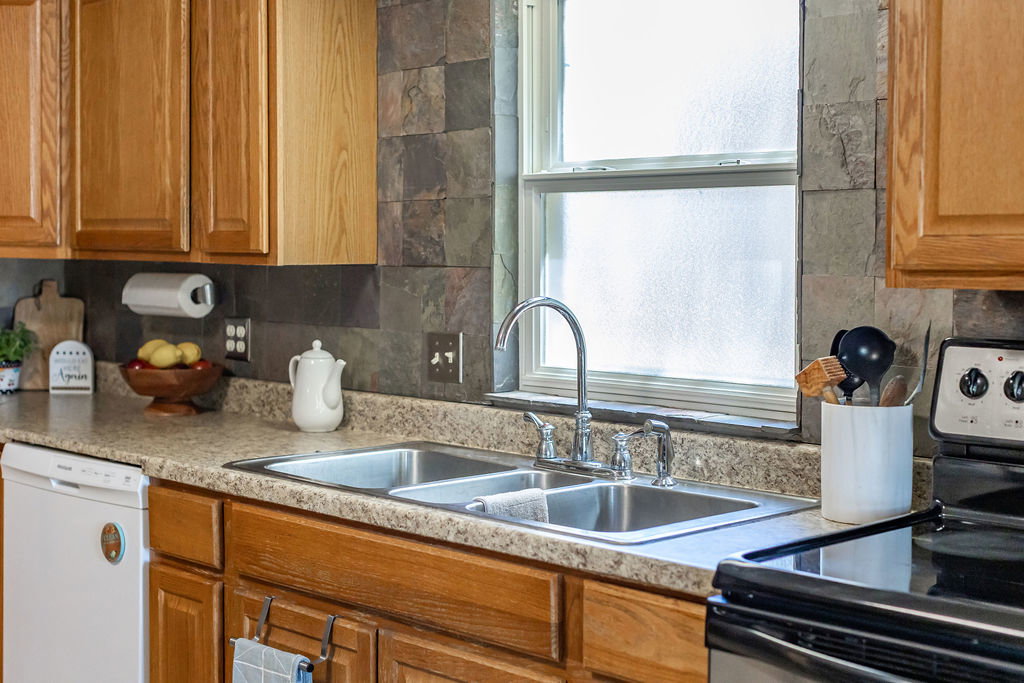 Kitchen featuring wood cabinetry, laminate countertops, stainless steel double basin sink with a high-arc faucet, and slate-style tile backsplash