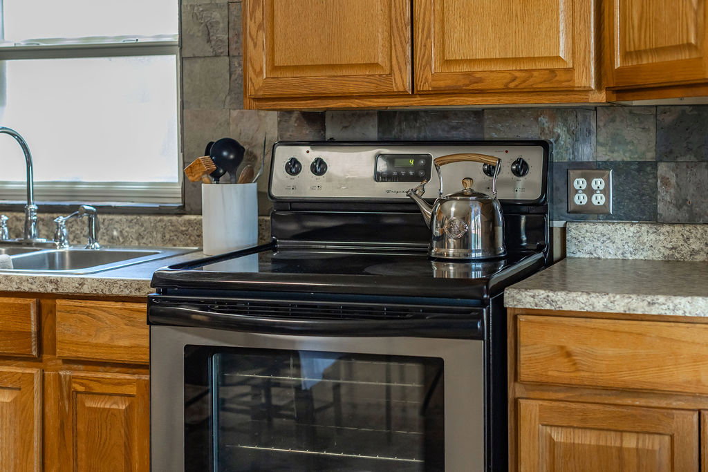 Stainless steel electric range with a visible oven, slate-style tile backsplash, wood-finish cabinetry, and speckled countertops