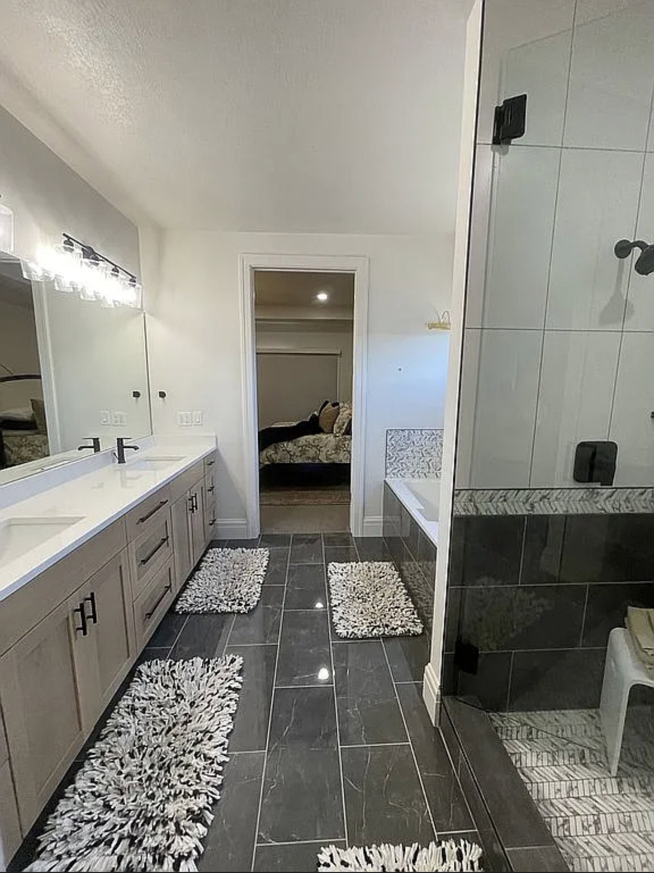 Bathroom featuring a double vanity with light wood-finish cabinetry and white countertops, a walk-in shower with glass enclosure and chevron pattern tile, a built-in tub with mosaic tile surround, and dark rectangular floor tiling