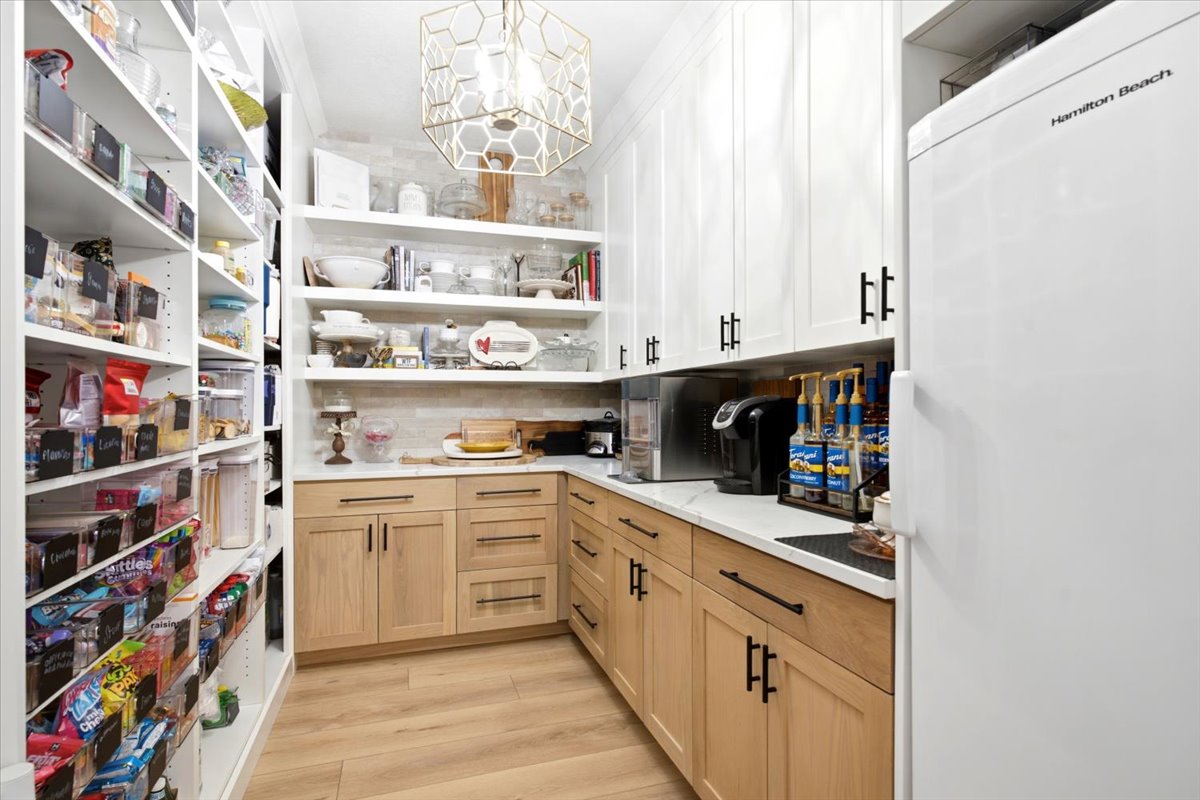 Custom pantry featuring extensive shelving, white upper cabinetry, natural wood-finish lower cabinetry, light stone-finish countertops, and a white refrigerator
