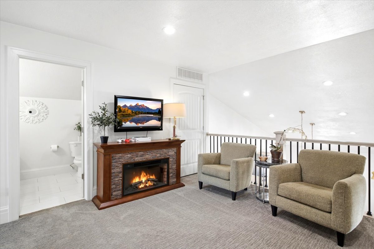 Carpeted loft area featuring a decorative stone-front electric fireplace with a wood-finish mantel, white walls, recessed lighting, and a black iron railing