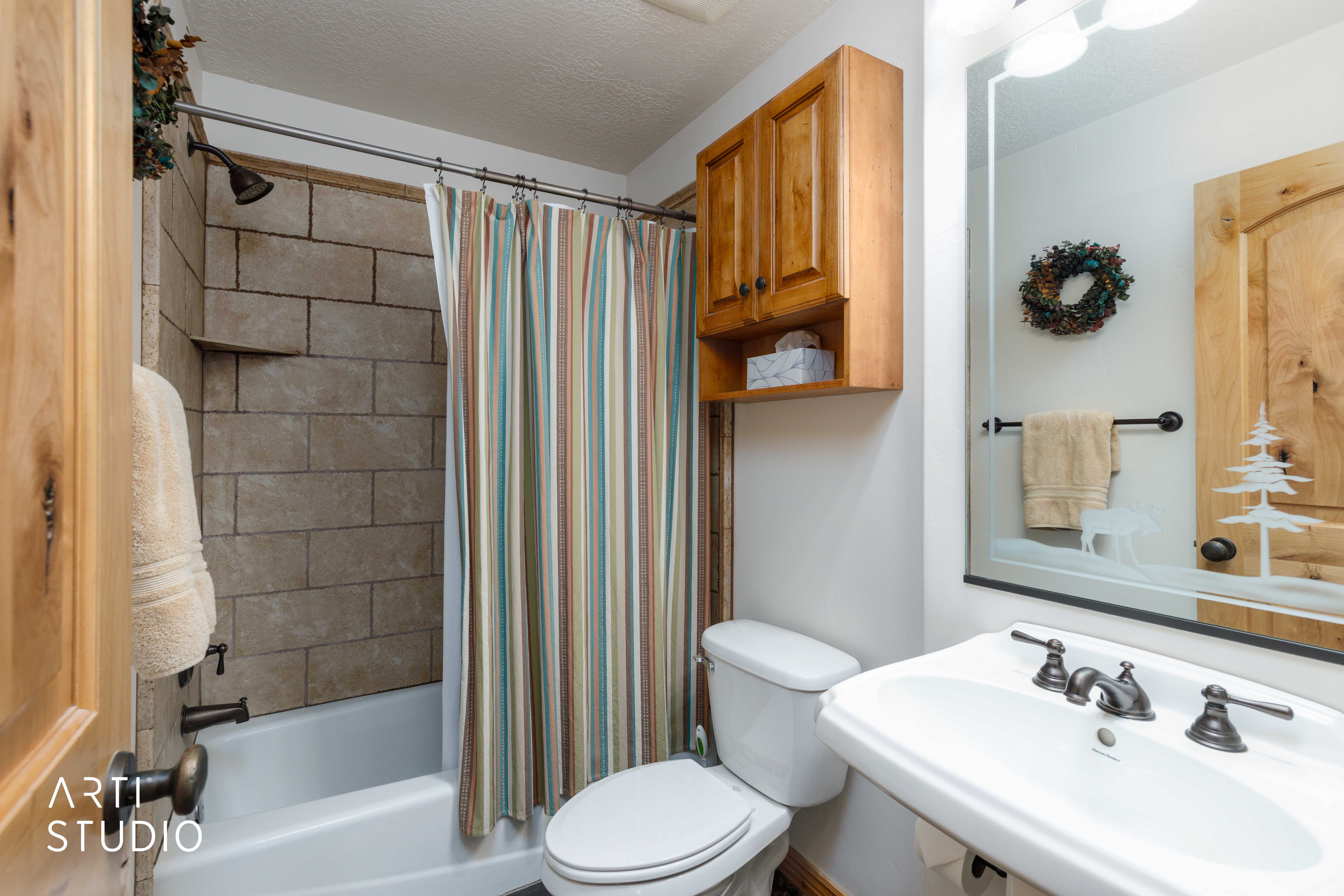 Full bathroom featuring a tub and shower combination with stone-look tile surround, a wall-mounted sink with a dark bronze-finish faucet, a toilet, a wood-finish wall cabinet, and a mirrored vanity with a decorative etched design