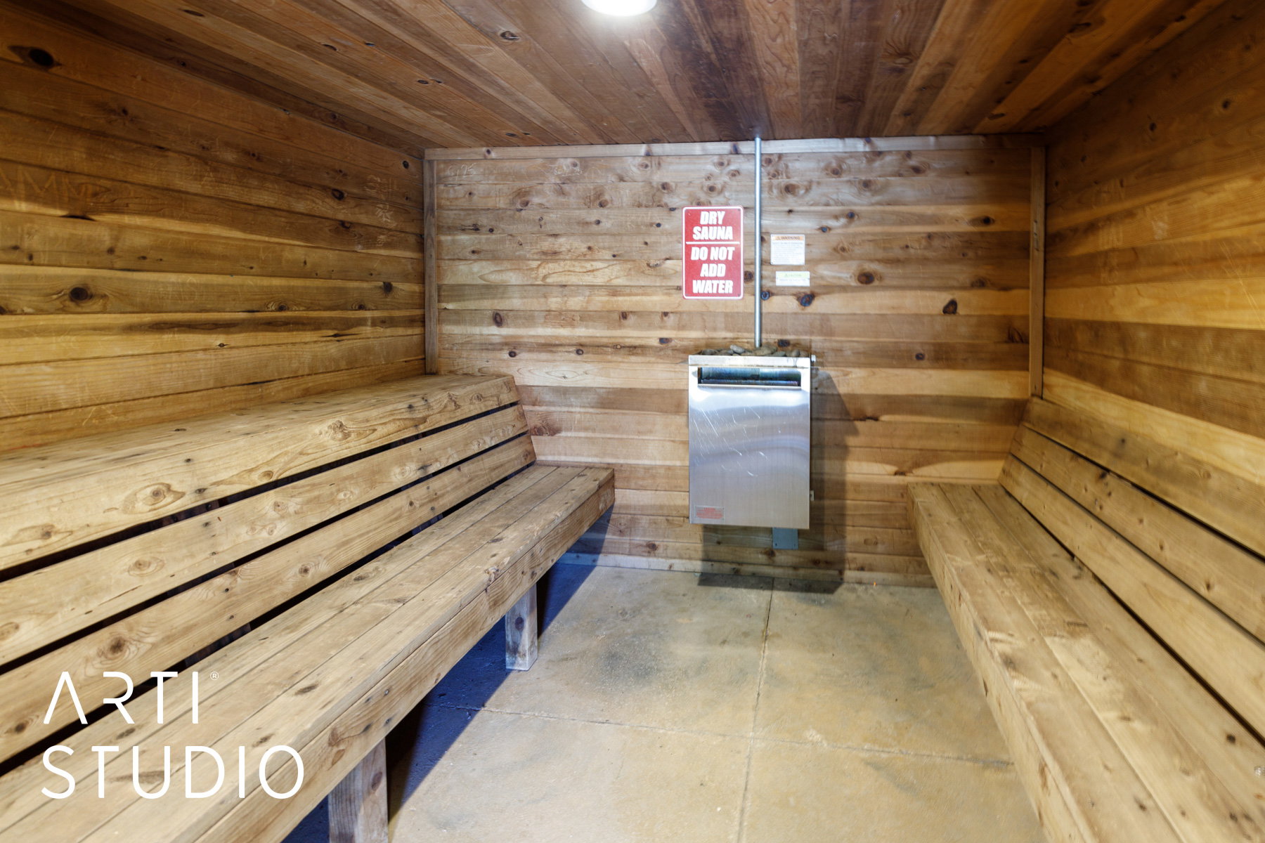 Sauna room featuring full wood paneling on walls and ceiling, two multi-tiered wooden benches, concrete flooring, and a wall-mounted stainless steel sauna heater