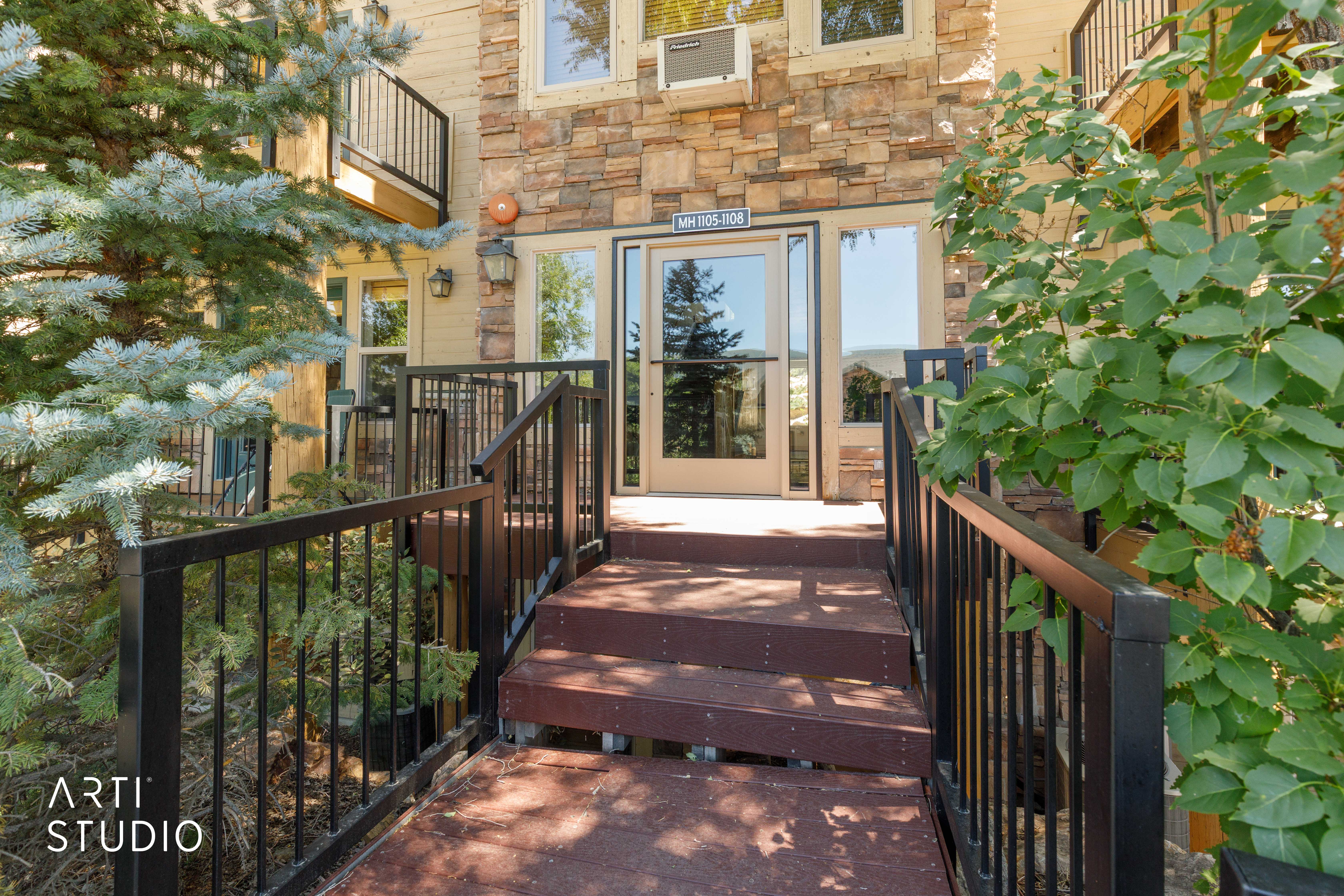 Exterior entrance featuring a stone facade, a raised wooden walkway, black metal railings, and a glass-paneled double door entry