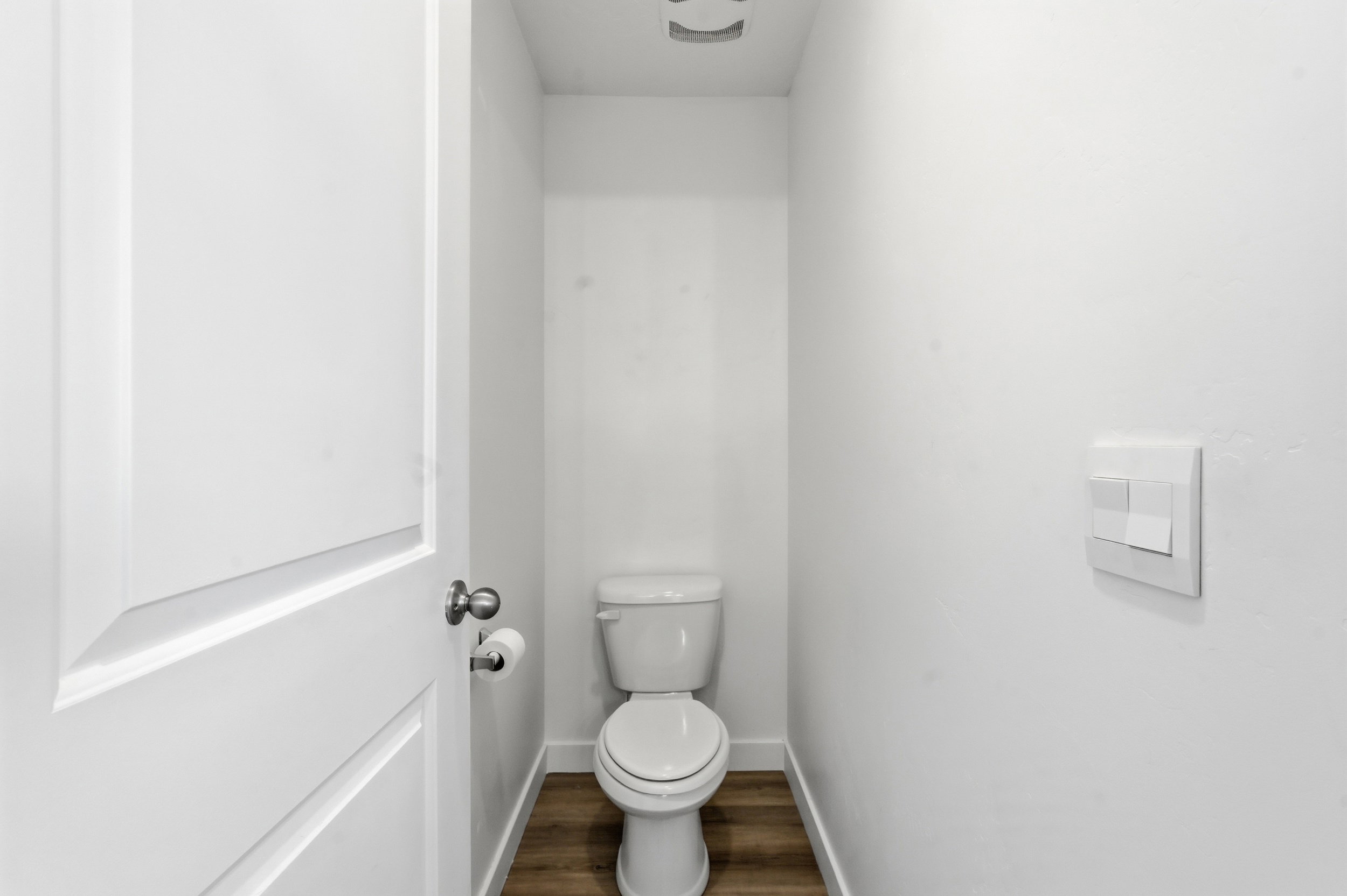 Compact water closet featuring a standard toilet, wood-finish flooring, white baseboards, and a contemporary wall switch