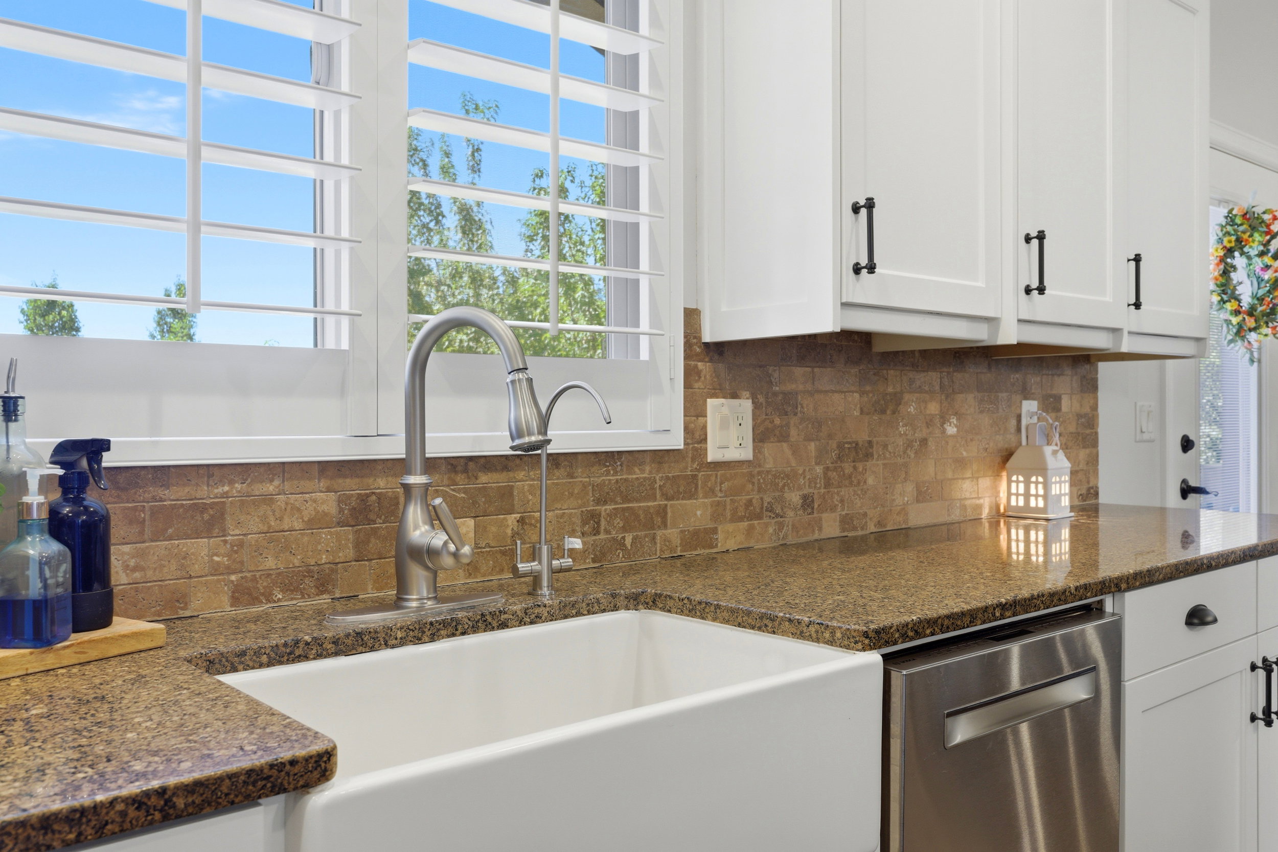 Deep farmhouse sink with a gooseneck faucet, plantation shutters, stone-finish subway tile backsplash, and dark granite countertops