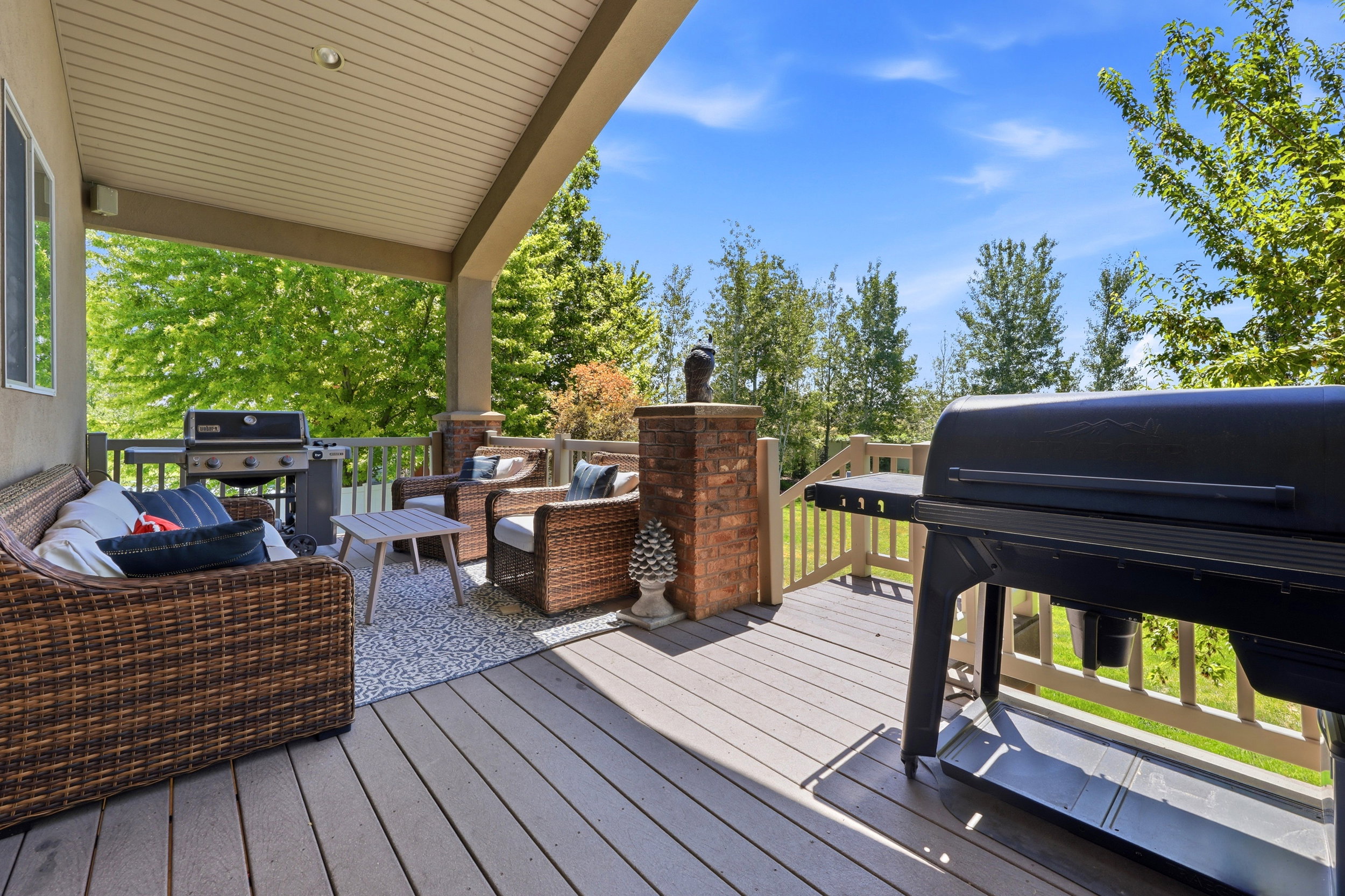 Spacious covered deck featuring composite decking, exposed aggregate flooring, brick pillar accents, and a wooden railing