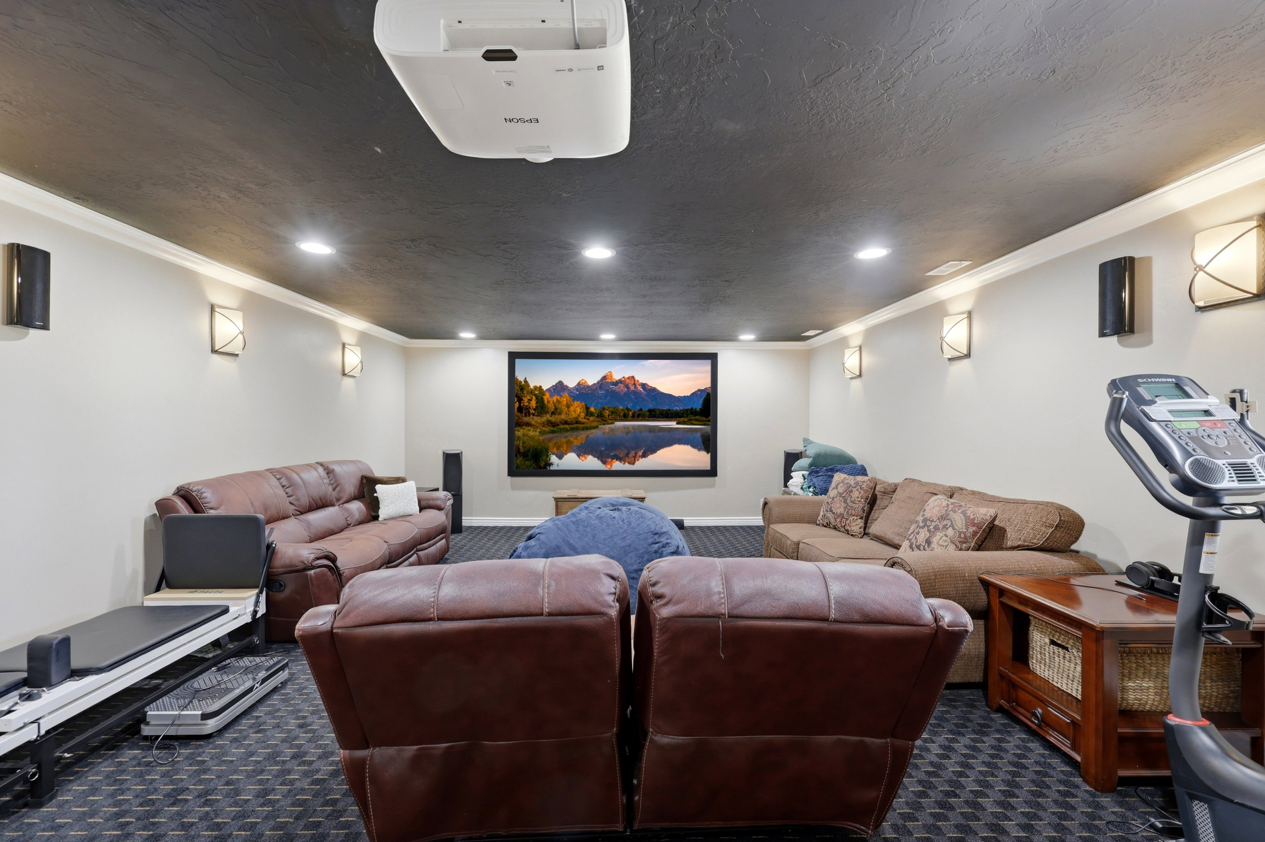 Spacious media room featuring a large projector screen, ceiling-mounted projector, recessed lighting, and wall sconces