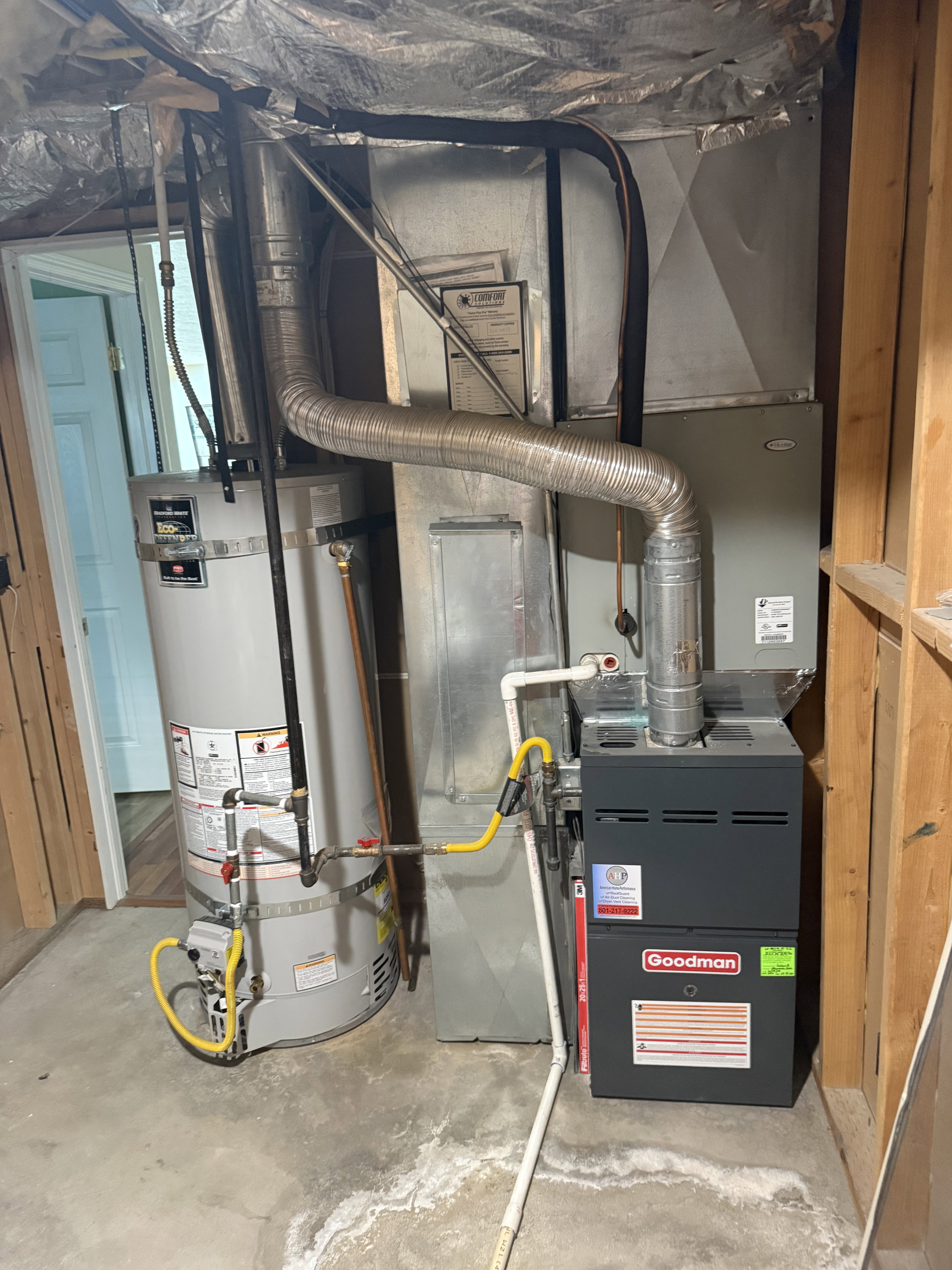 Goodman brand furnace and water heater installation with visible ductwork, gas lines, and PVC piping on a concrete floor