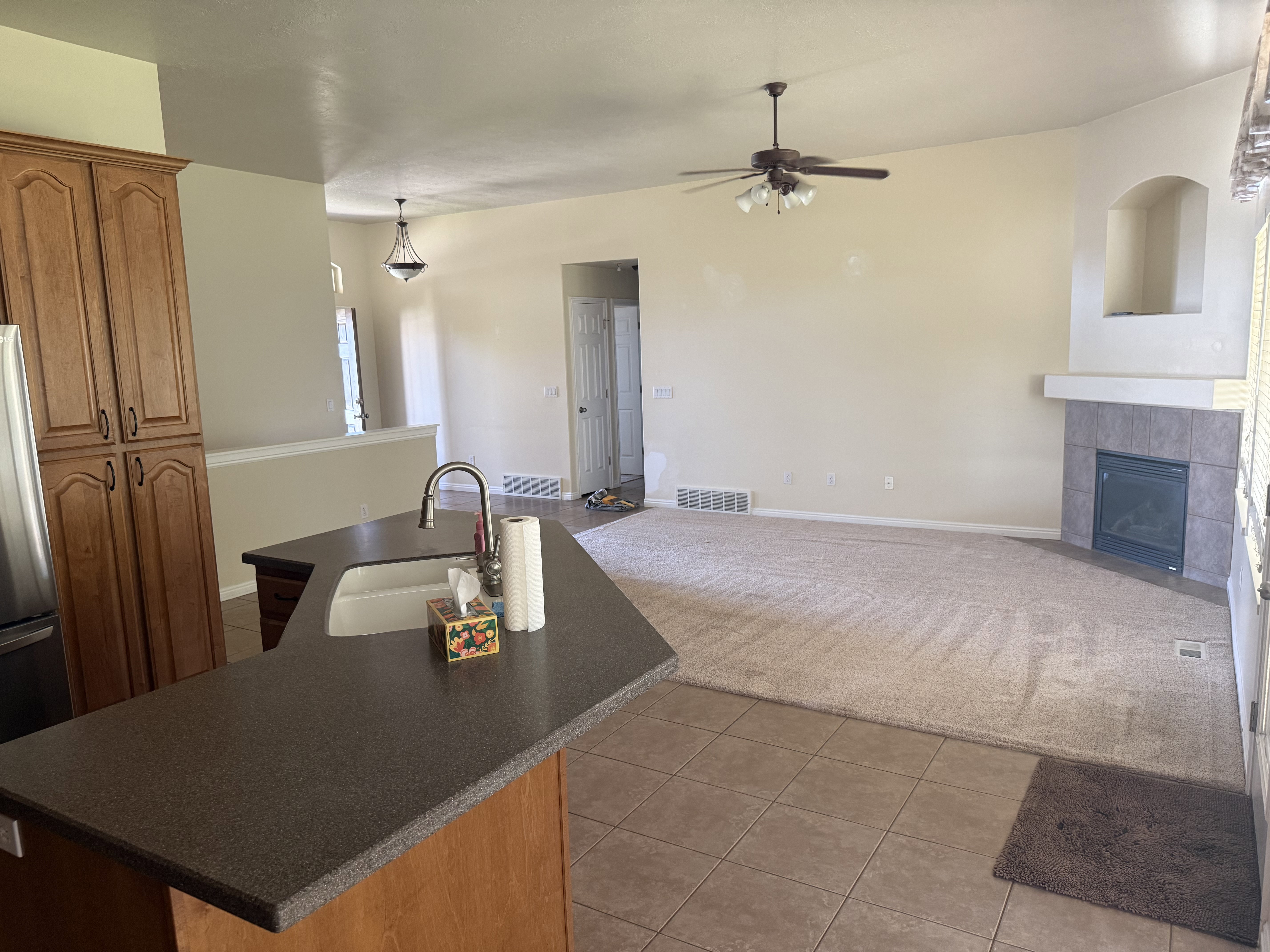 Open-concept living area featuring a kitchen island with a double basin sink, ample wood cabinetry, a ceiling fan with integrated lighting, a gas fireplace with a tiled surround, and a decorative arched niche