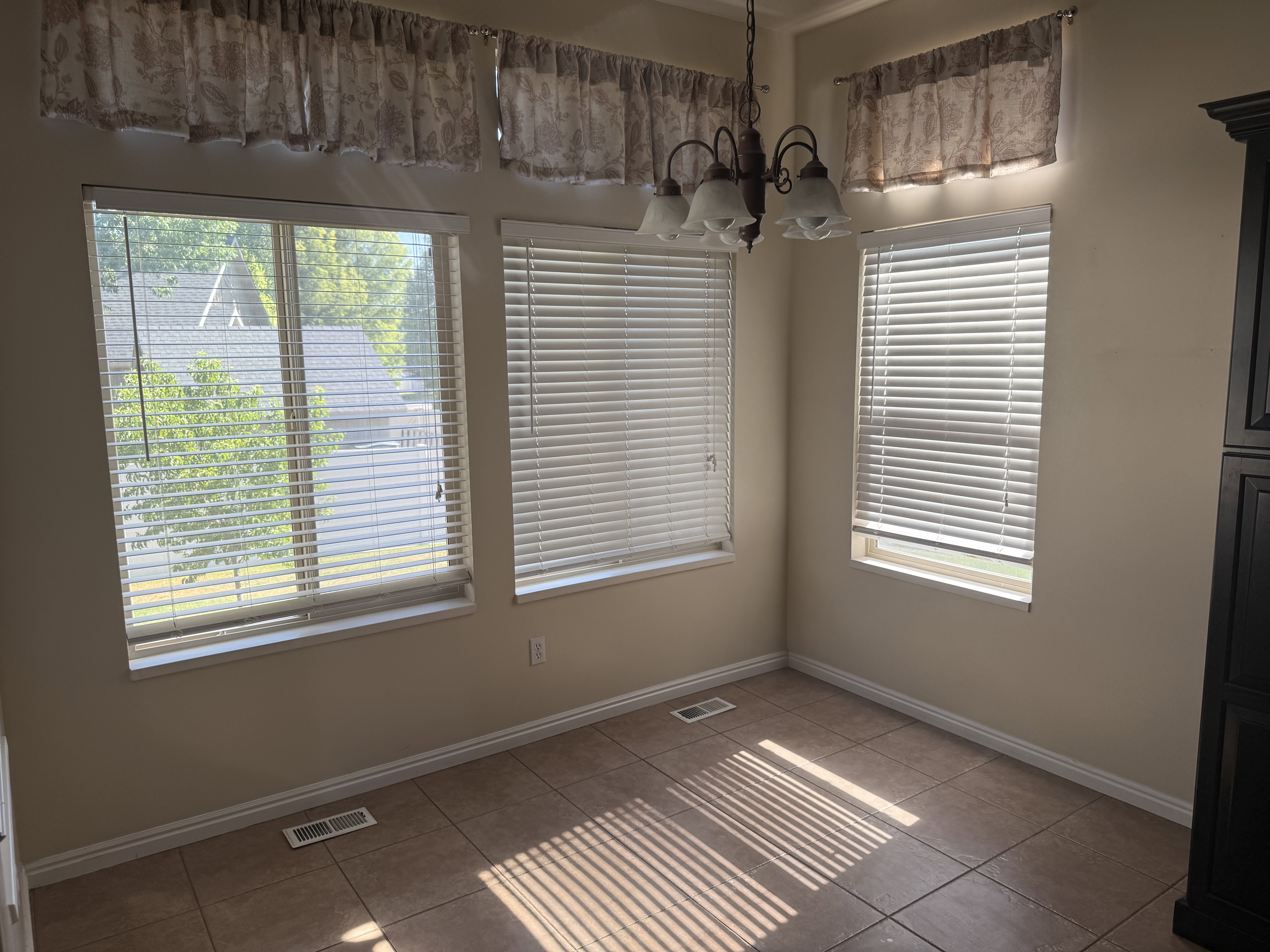 Room featuring three windows with horizontal blinds, a ceiling-mounted light fixture, tile flooring, and a built-in dark wood cabinet