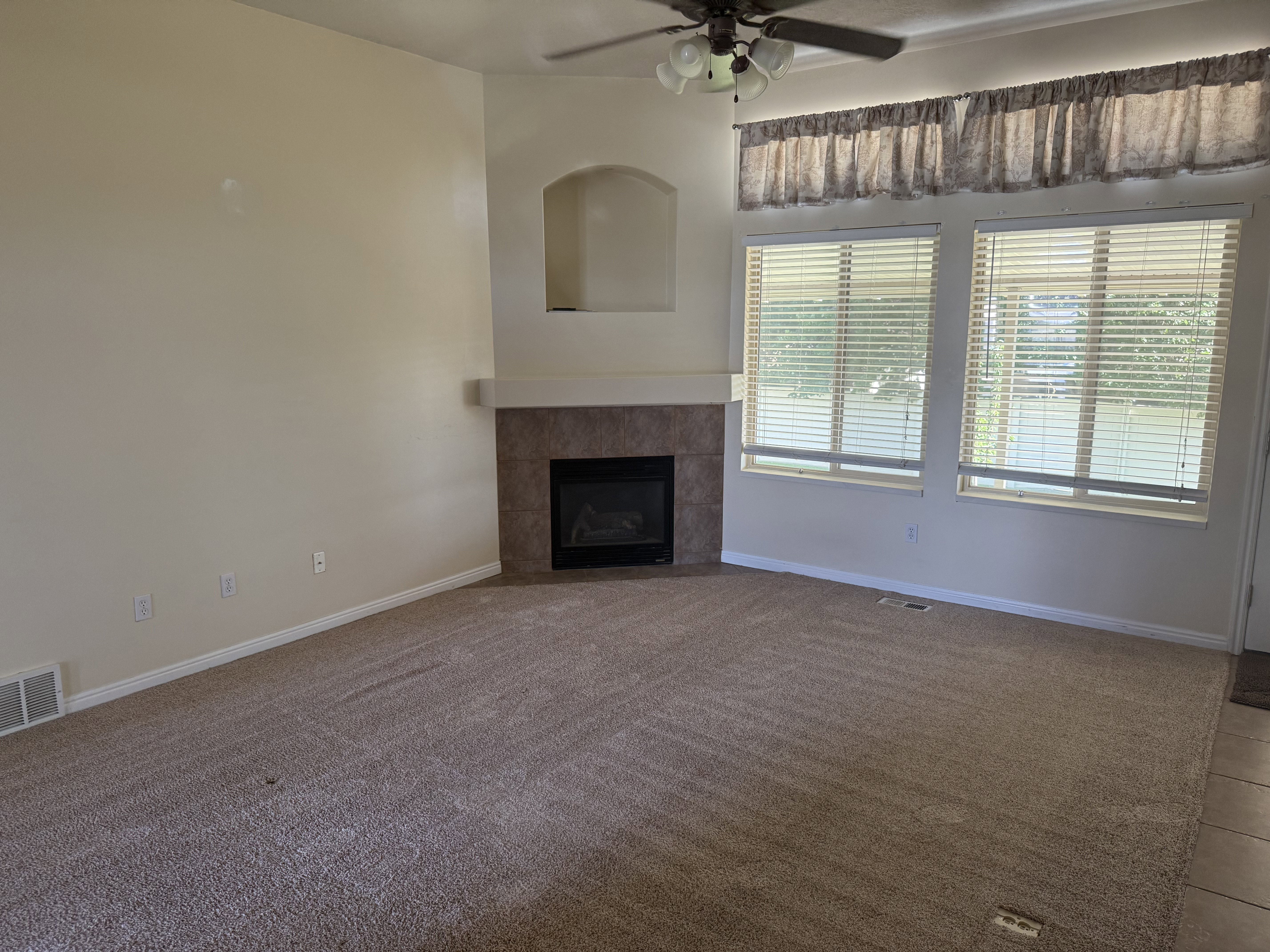 Living area featuring an integrated fireplace with a tile surround and mantel, two large windows with horizontal blinds, a ceiling fan with integrated lighting, and a neutral carpet