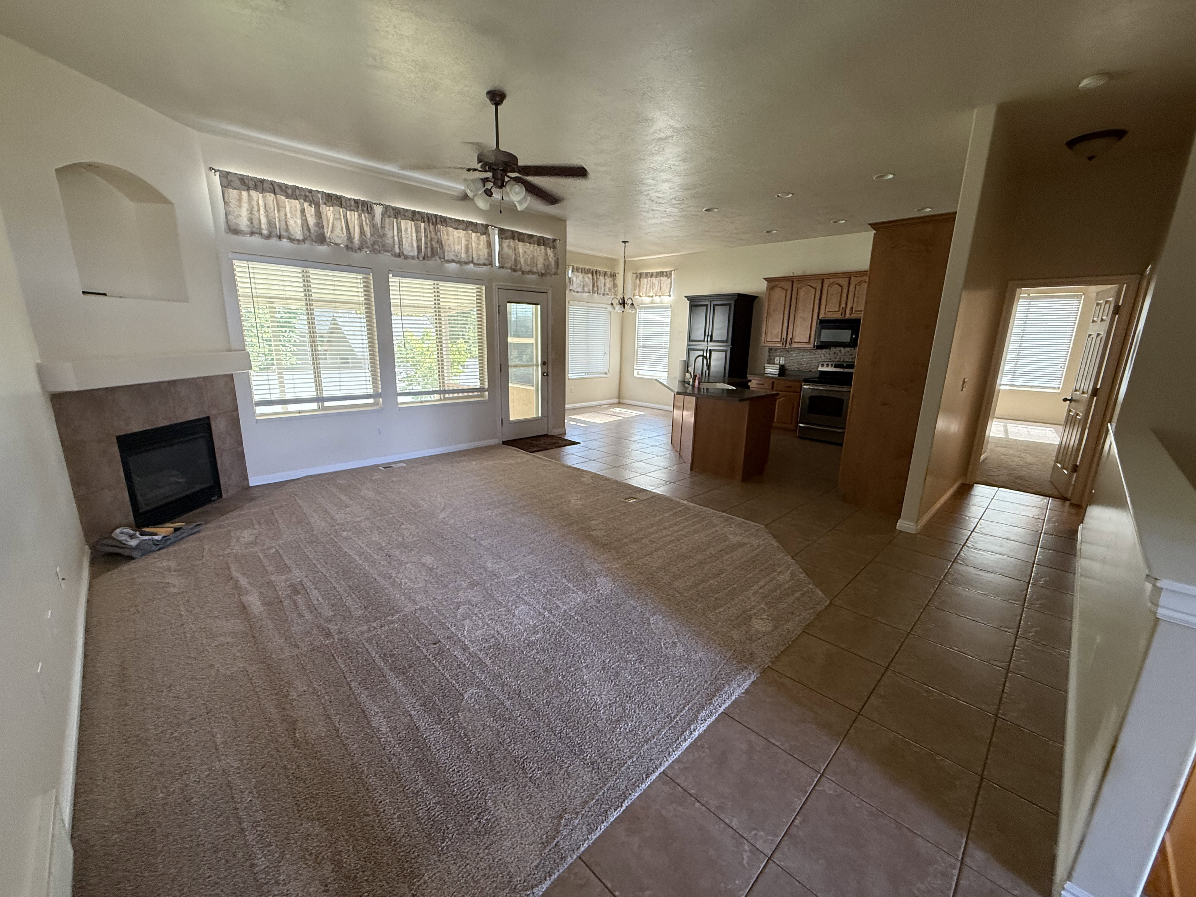 Open-concept living space featuring a fireplace with a tiled surround and mantel, expansive windows, a ceiling fan, and a kitchen with wood cabinetry and tile flooring